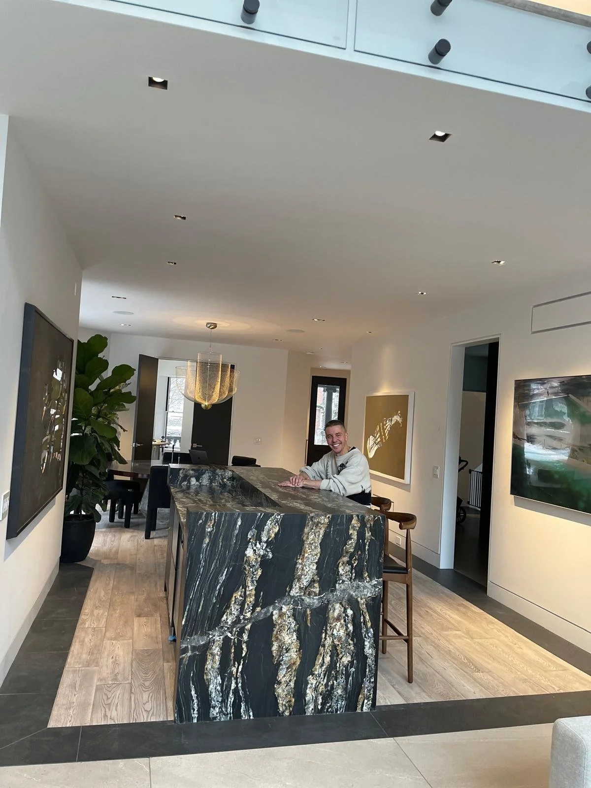 A happy customer is sitting at a kitchen island with a black and gold marble pattern in a modern, well-lit home. The kitchen features surrounding artwork and a large potted plant, with visible dining area and windows in the background.