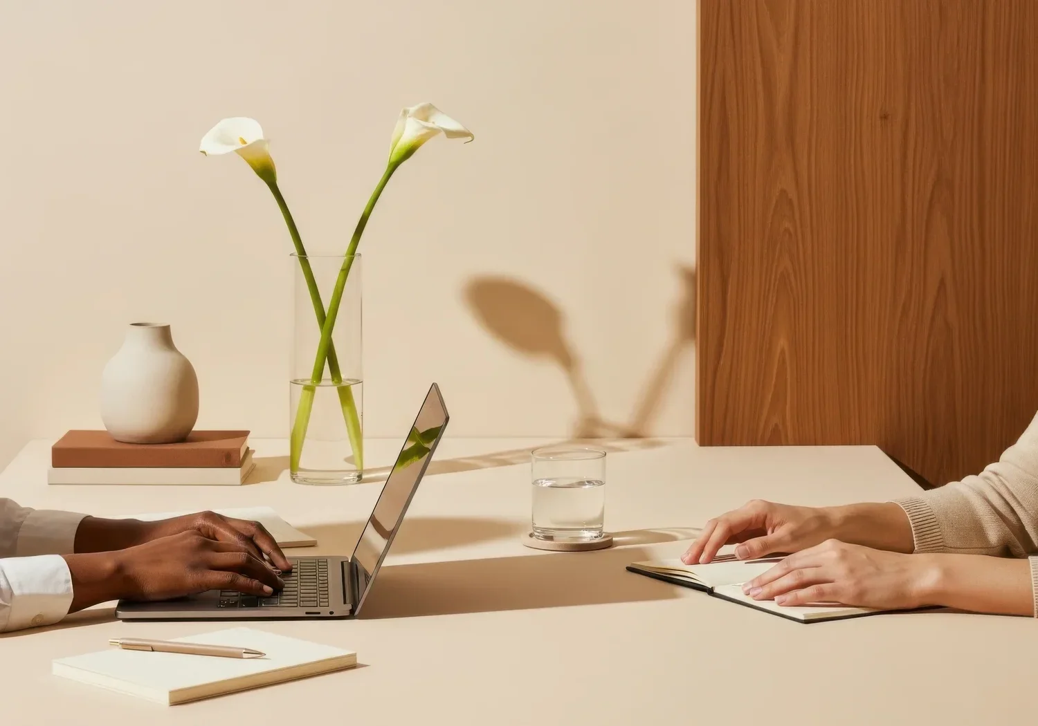 Two people sitting across from each other at a table, one person using a laptop and the other taking notes, with a vase holding two calla lilies and a glass of water on the table.