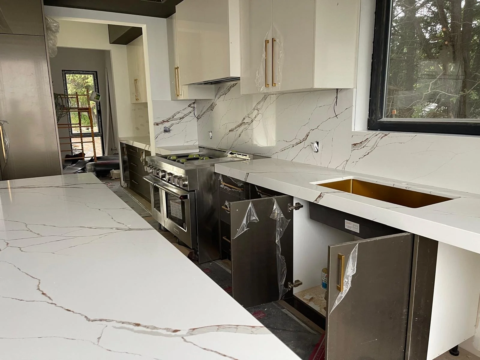New kitchen under installation construction with white marble countertops and backsplash, black and gold cabinet handles, stainless steel stove, and an unfinished sink area.