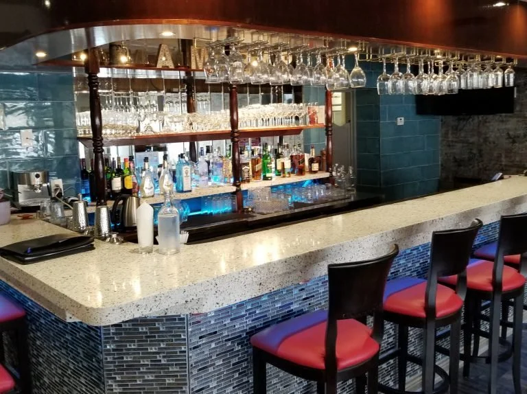 A marble bar with a curved cream-colored countertop, dark tile base, and a glass shelf behind stocked with liquor bottles and hanging wine glasses.