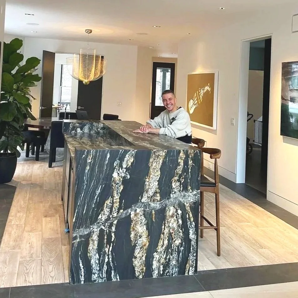 A happy customer sitting at a large, polished marble kitchen island with a dark and gold veined pattern, smiling and leaning on the counter. The kitchen has light wooden flooring, a modern chandelier, and artwork on the walls.