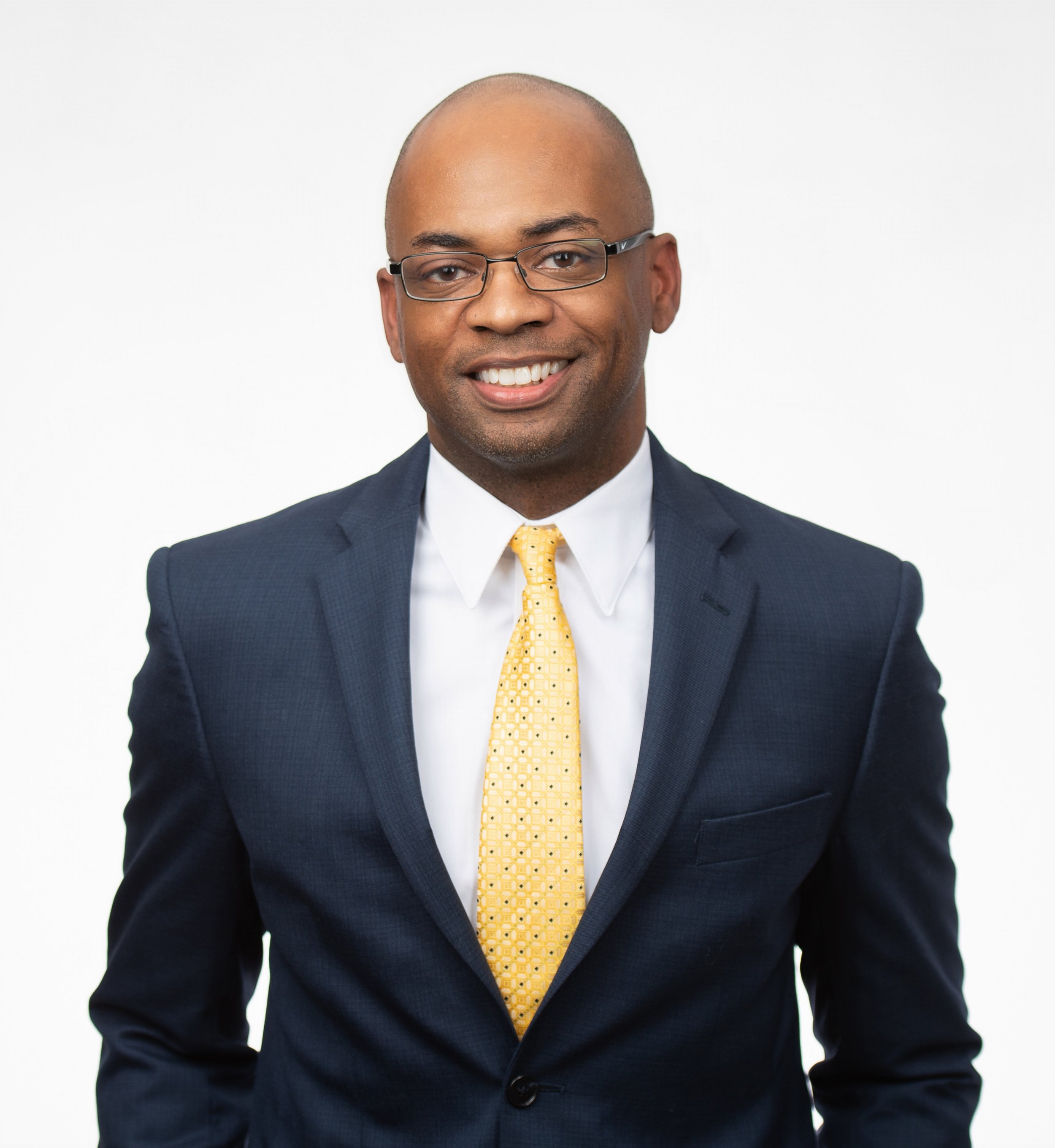 Man in a dark suit with a yellow tie and glasses, smiling, on a white background.
