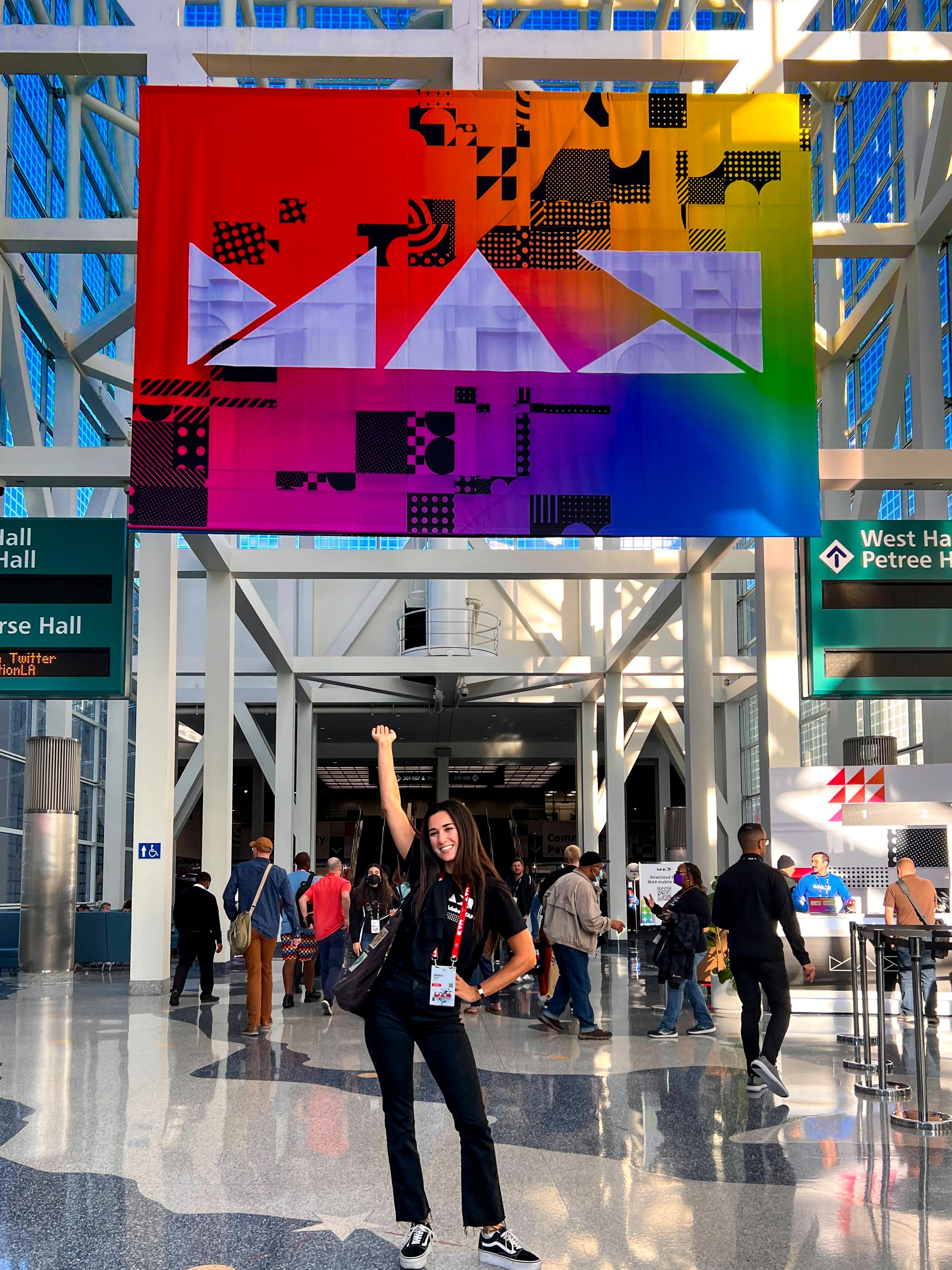 A vibrant event hall with a large, multicolored banner displaying abstract geometric designs hung above. People are walking around, with one person in the foreground posing cheerfully. Signage directs towards West Hall and Petree Hall. The atmosphere