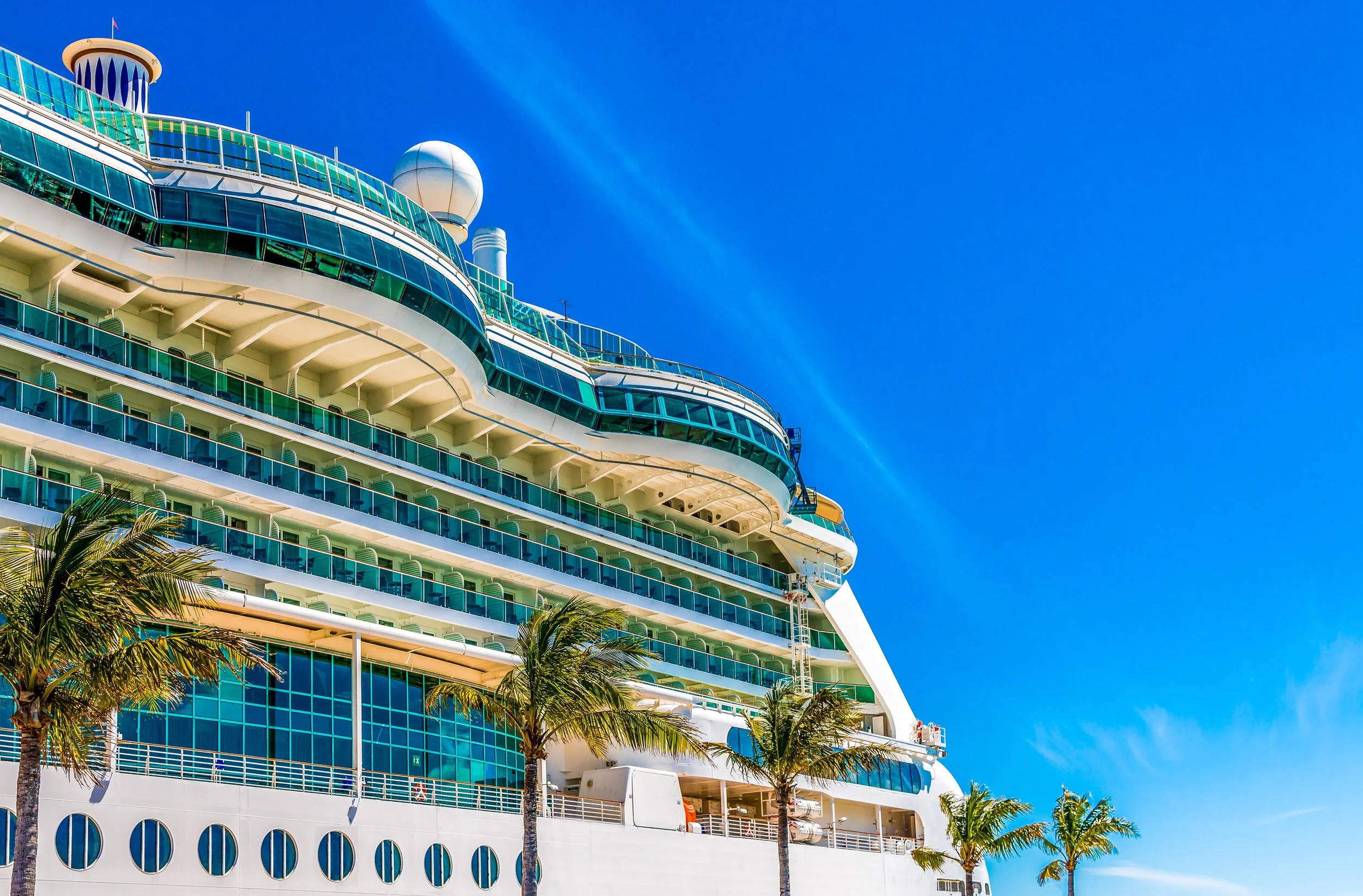 A large cruise ship with multiple decks, glass windows, and balconies, docked near palm trees against a bright blue sky.