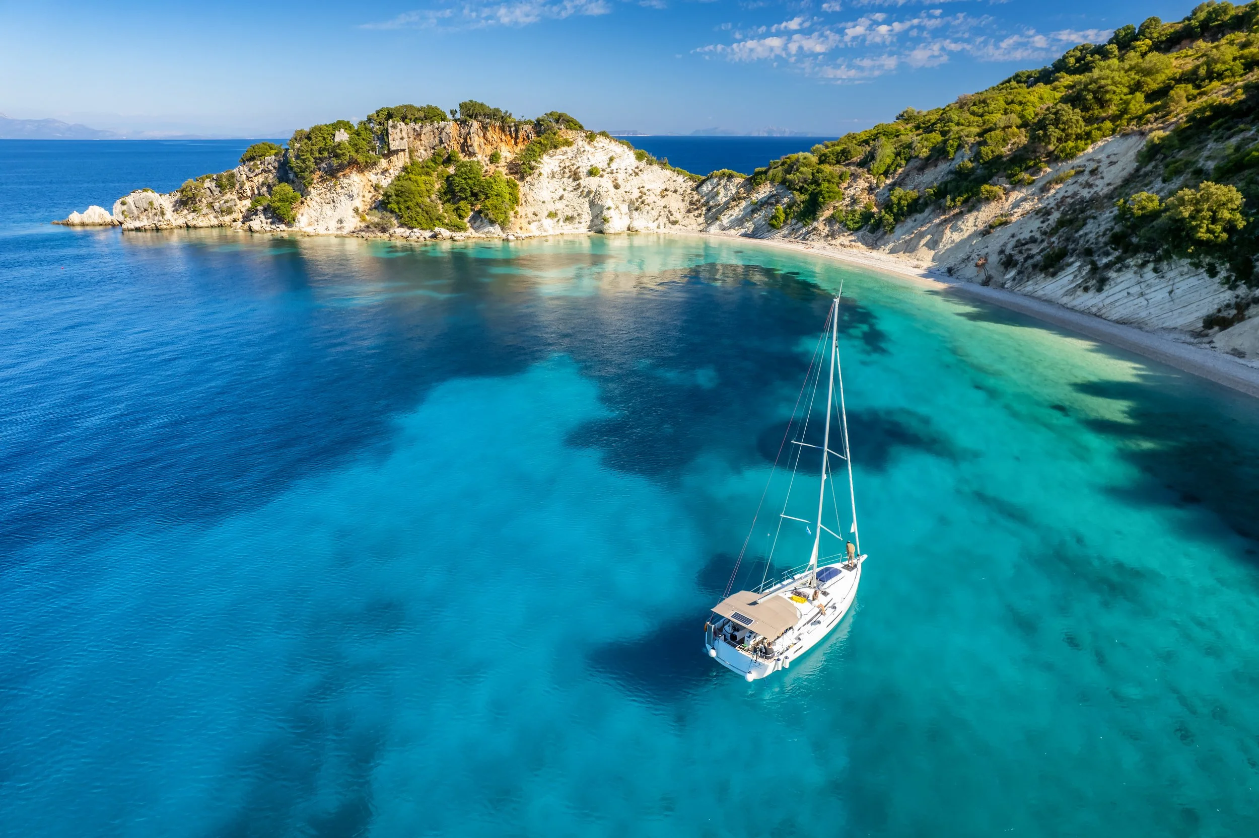 A white sailboat with beige canopy floating in clear blue water near a green, rocky shoreline with trees, under a partly cloudy sky.