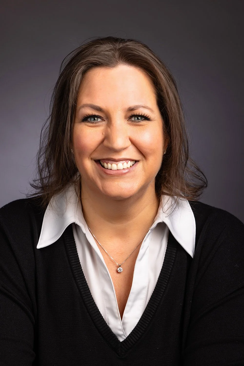 A smiling woman with shoulder-length brown hair, wearing a white collared shirt and black sweater, against a dark gray background.