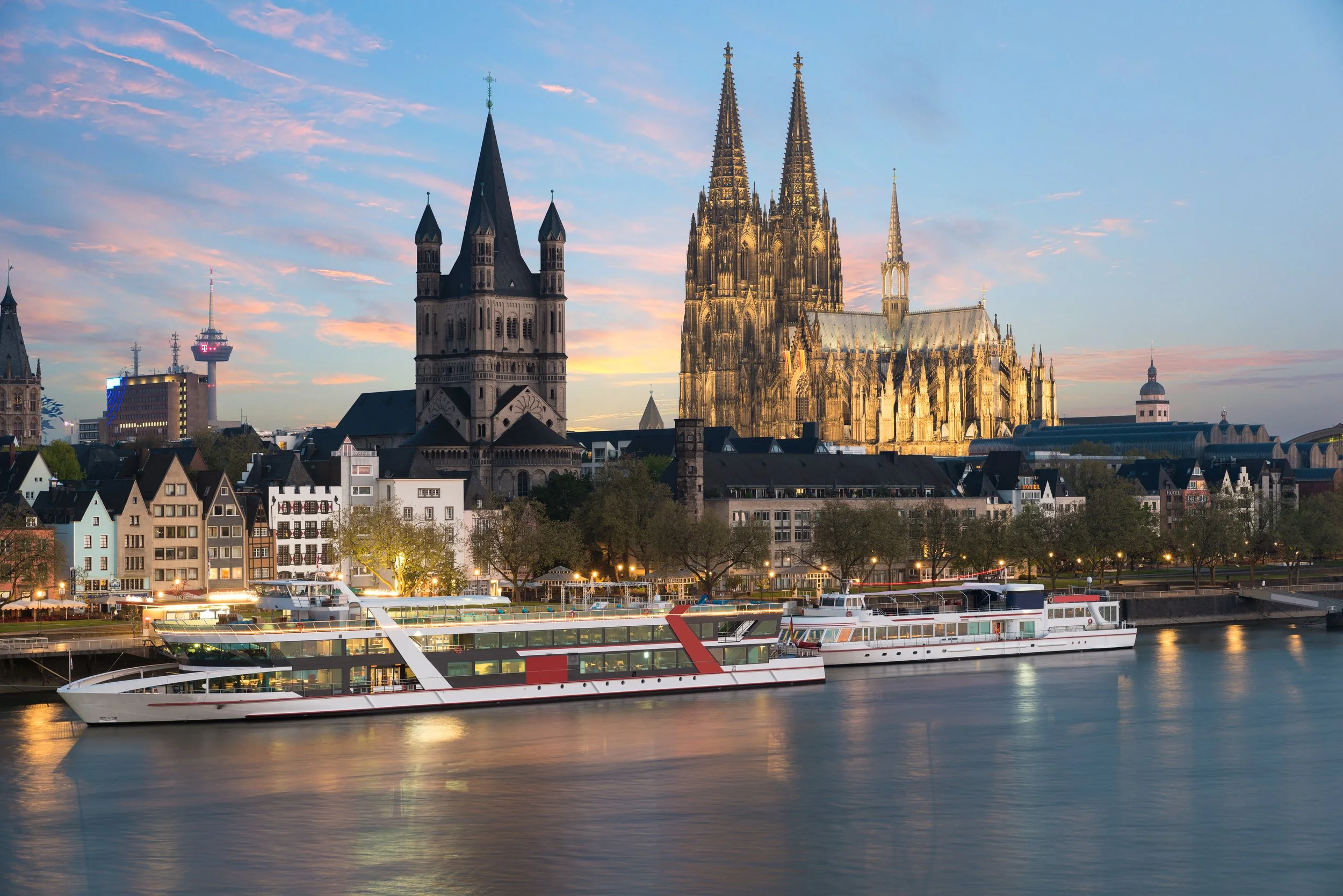Sunset view of Cologne, Germany, with the Cologne Cathedral and Old Town buildings along the Rhine River, boats on the water, and a partly cloudy sky.