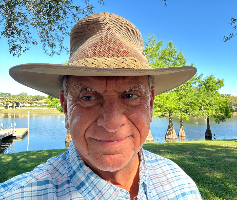 Mitch Baker wearing a wide-brimmed straw hat and a checkered shirt is outdoors near a lake with trees and clear blue sky in the background.