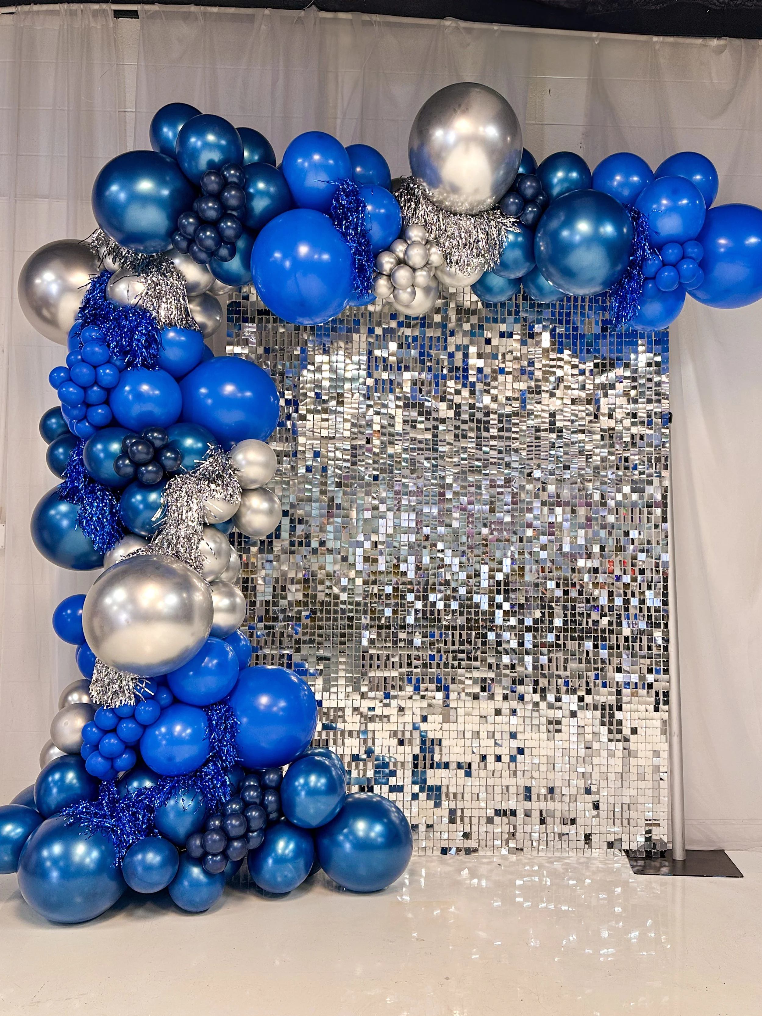 Blue, silver, and navy balloon arch with metallic and glitter accents in front of a silver mirrored backdrop.