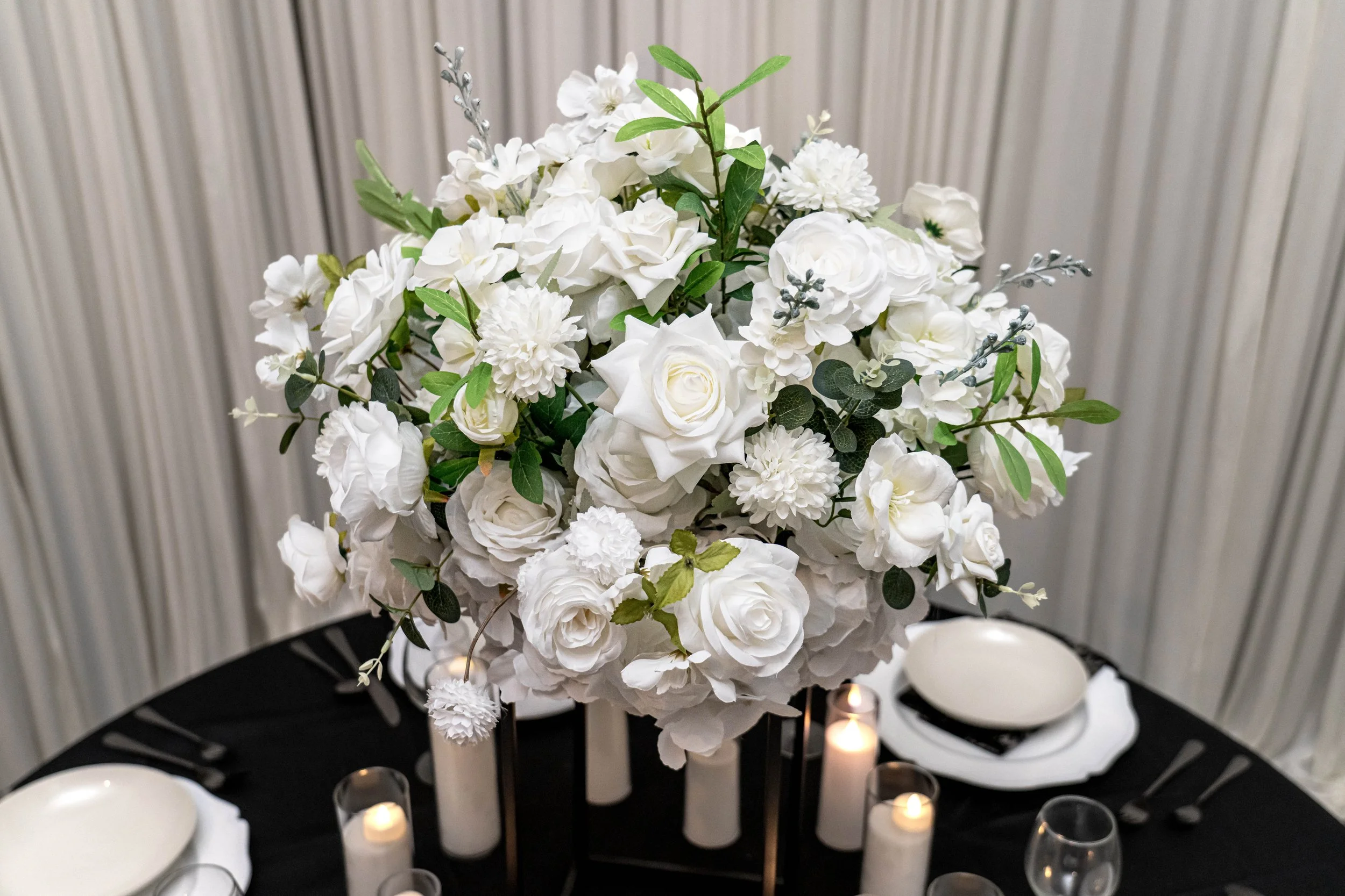 A floral centerpiece with white flowers and green leaves on a black table, surrounded by lit candles and decorative bowls.