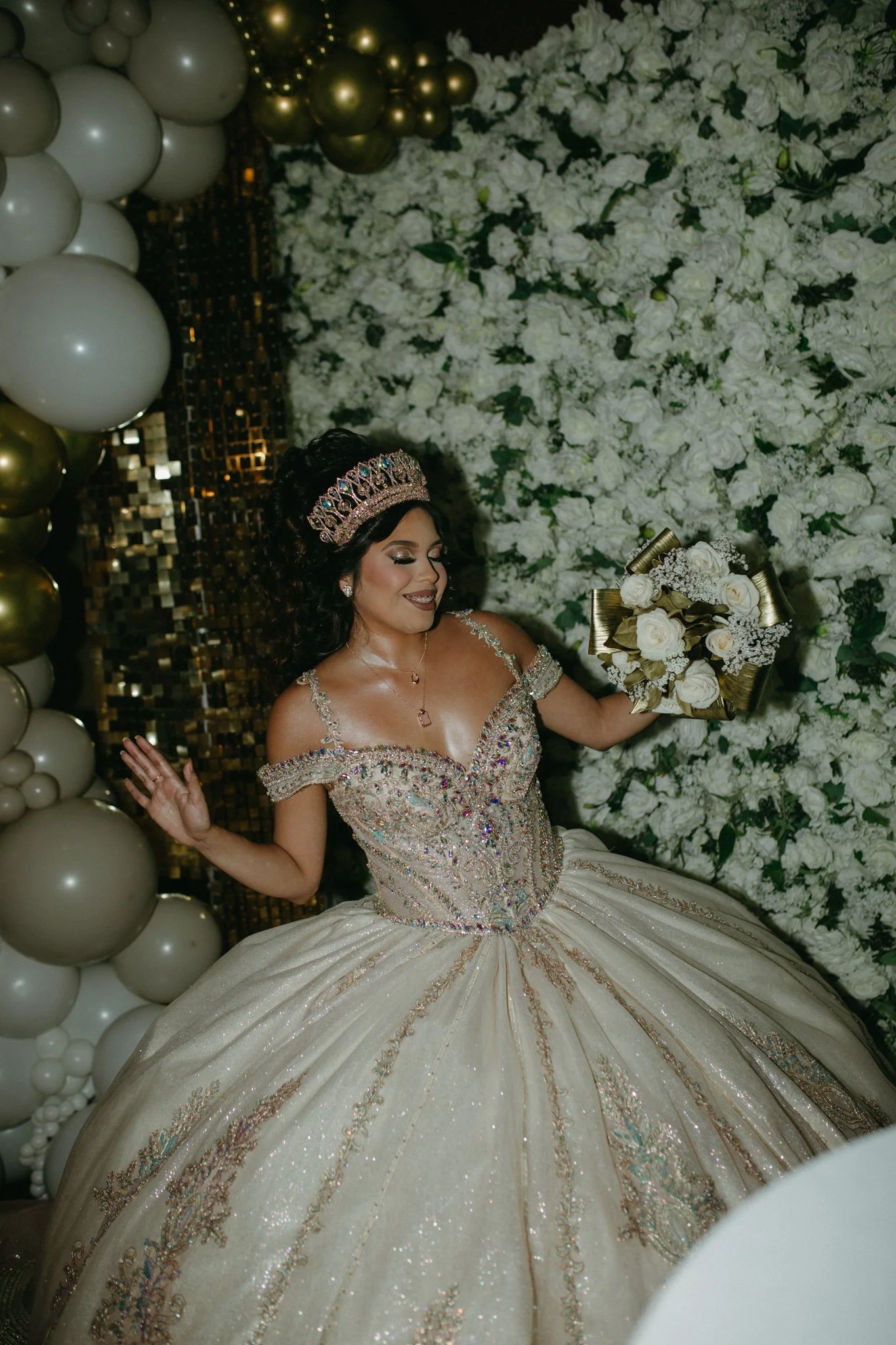 A bride in an ornate off-the-shoulder wedding gown with intricate beadwork and embroidery, holding a bouquet of white roses, posing in front of a wall of white flowers and surrounded by gold and white balloons.