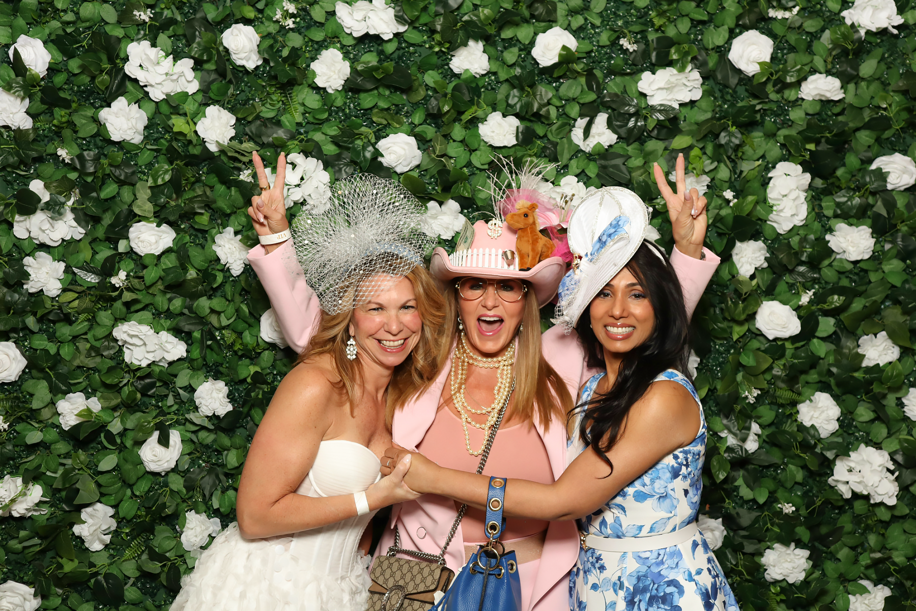 Three women smiling and celebrating at a photo booth, wearing elaborate hats and dresses, with a green floral backdrop.