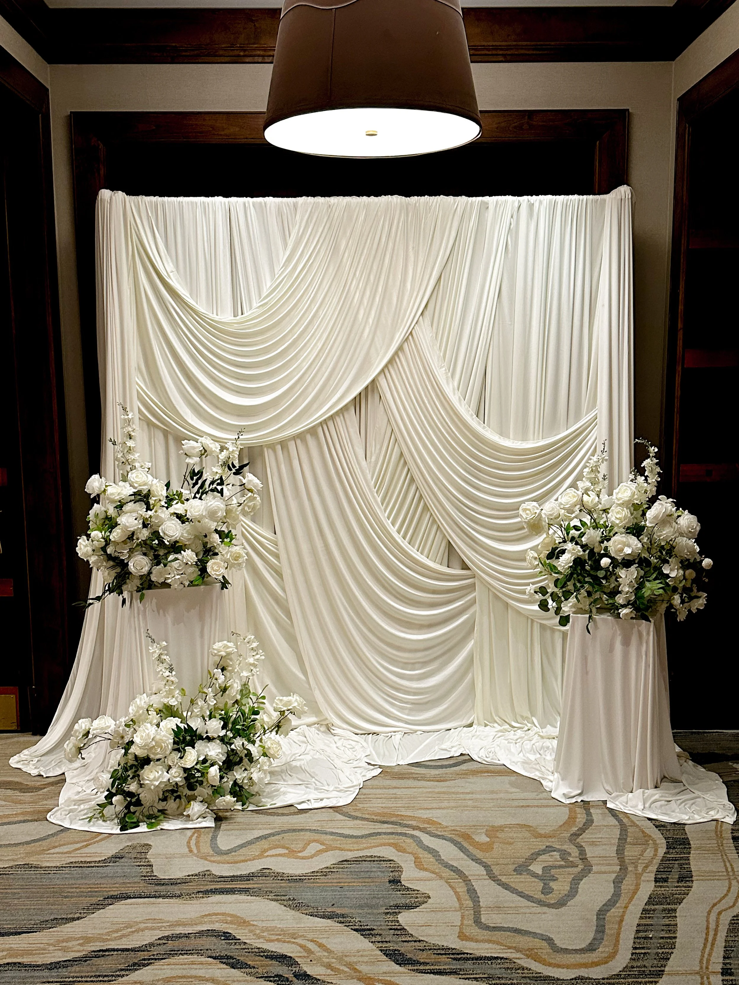 White wedding backdrop with draped fabric, floral arrangements of white roses at the base, and a dark brown lampshade hanging from the ceiling.