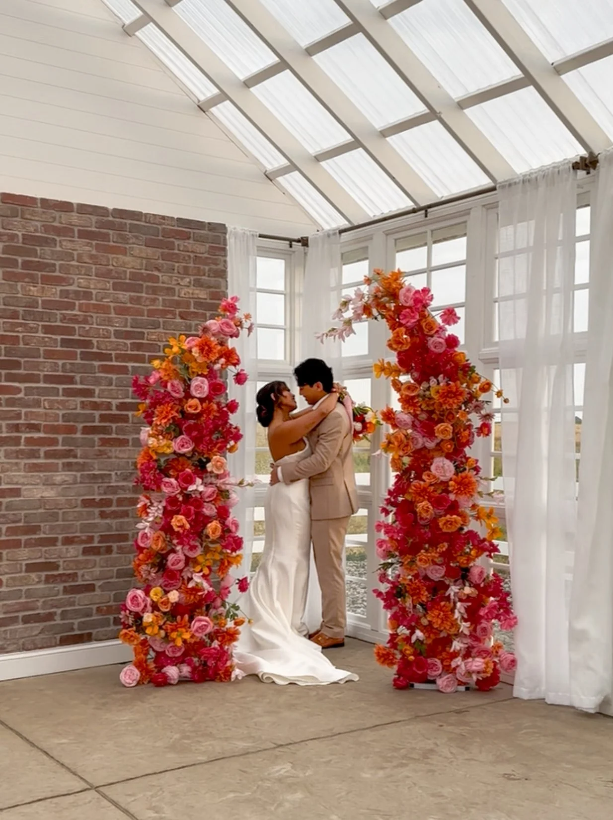 A couple sharing a kiss during their wedding ceremony, standing in front of a floral arch decorated with pink and orange flowers inside a room with large windows, white curtains, brick and white walls, and a glass ceiling.