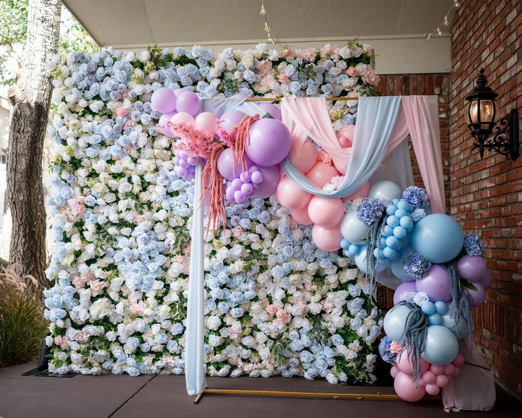 Floral backdrop decorated with pastel-colored balloons, pink and blue drapes, and pink tassels, likely set up for a celebration or wedding.