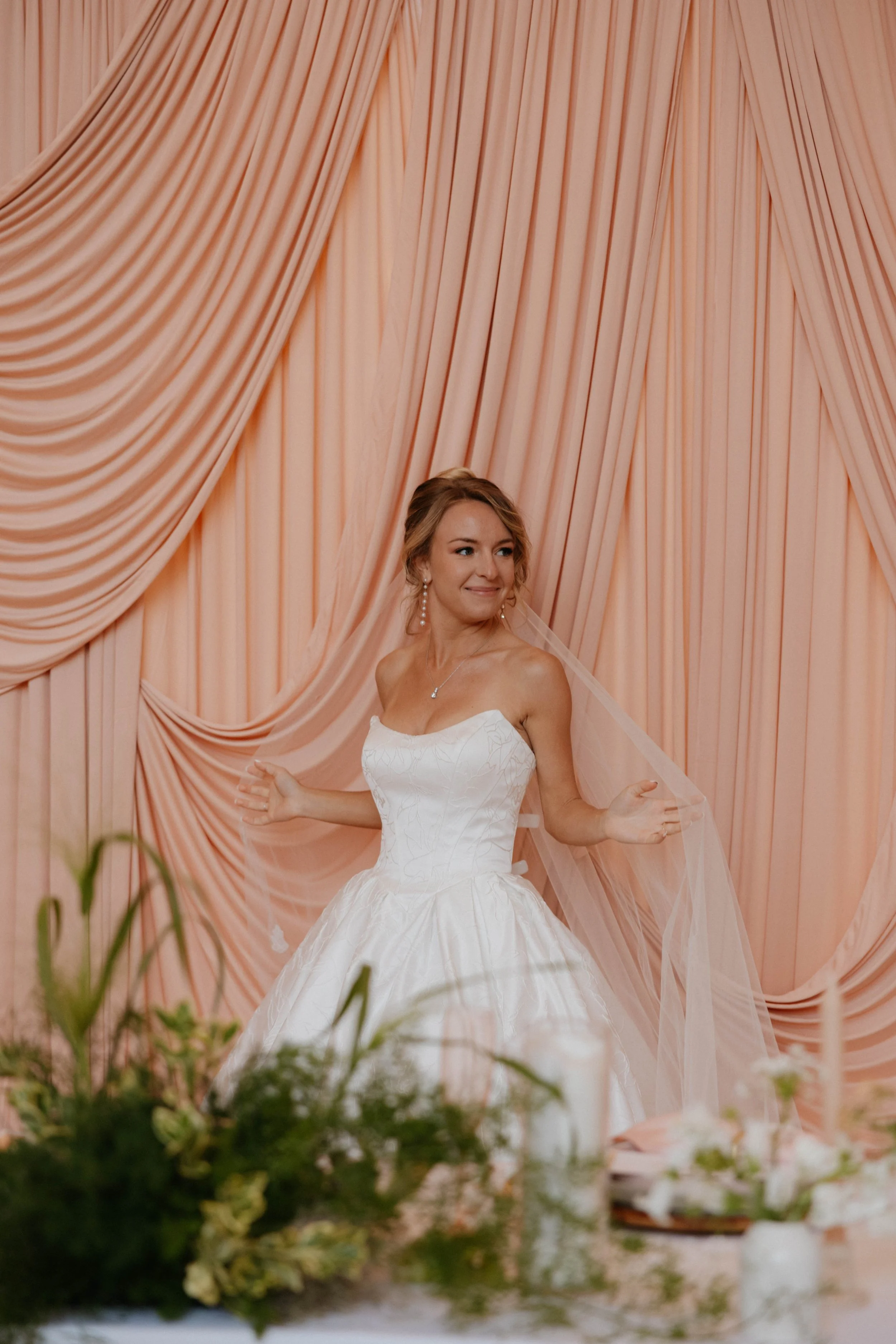 A bride in a white wedding gown with a strapless bodice and full skirt, smiling and posing in front of peach-colored draped curtains, with floral arrangements in the foreground.