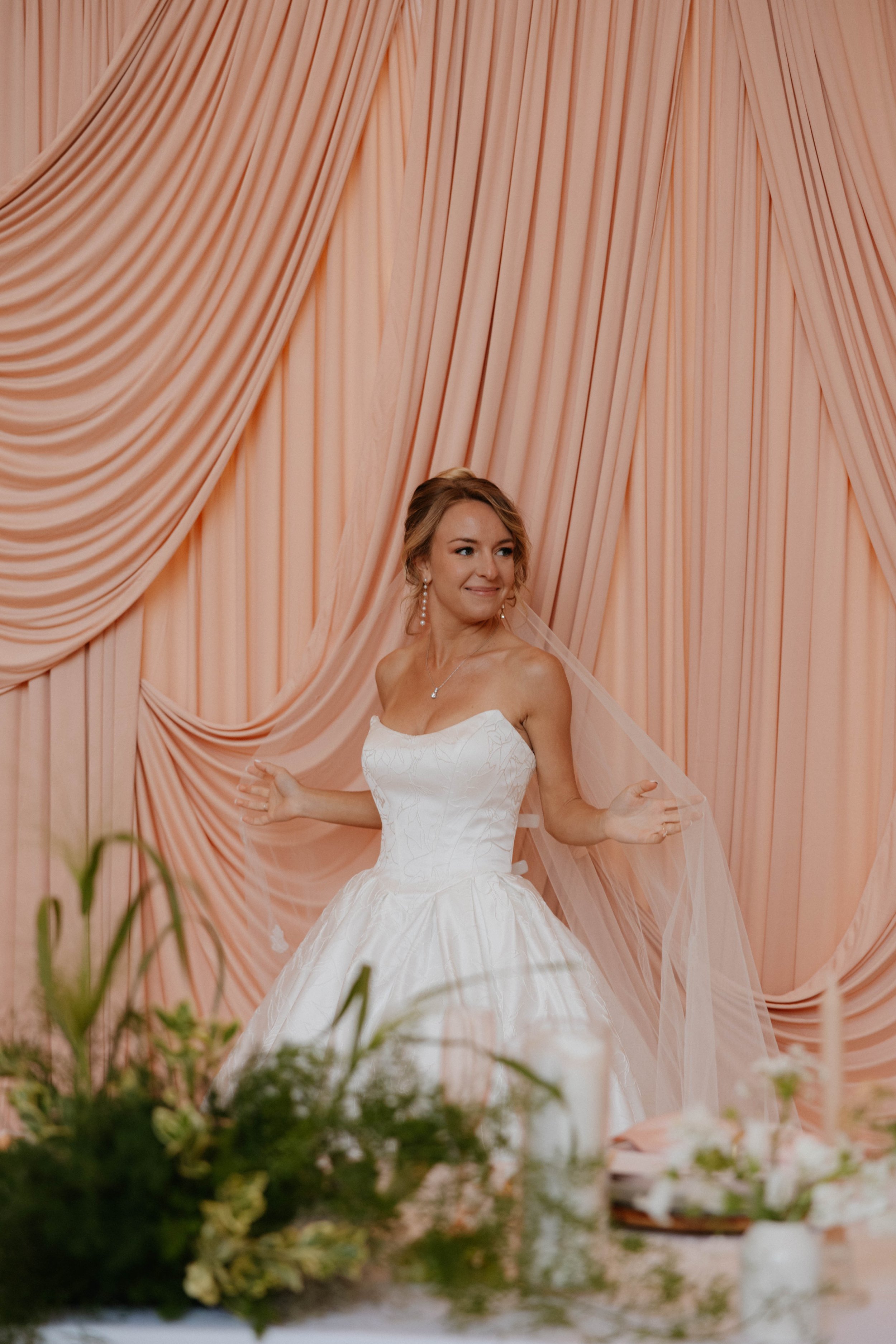 A bride in a white wedding gown with a strapless bodice and full skirt, smiling and posing in front of peach-colored draped curtains, with floral arrangements in the foreground.