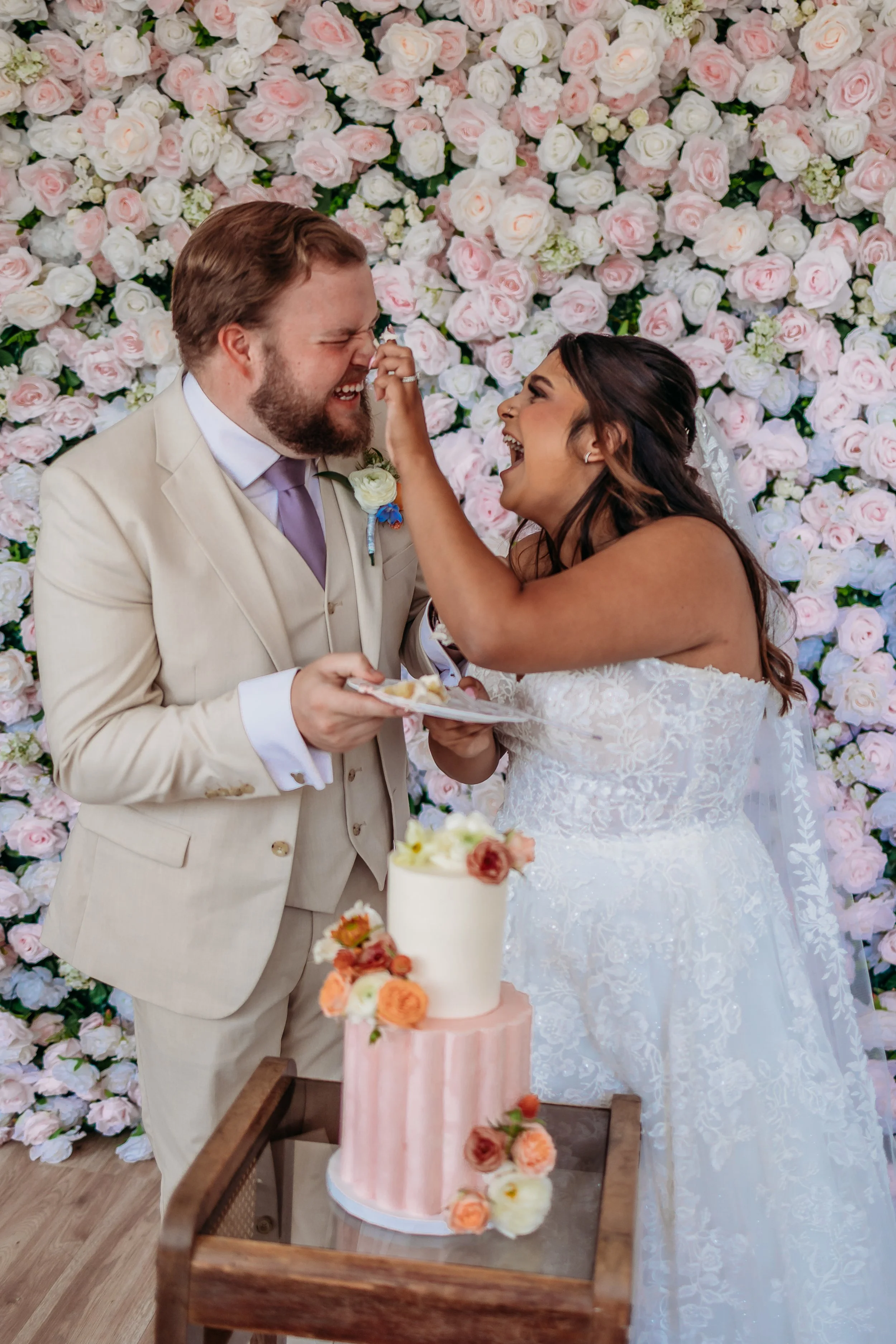 A bride and groom sharing a joyful moment at their wedding with a floral backdrop and a decorated cake in front of them.