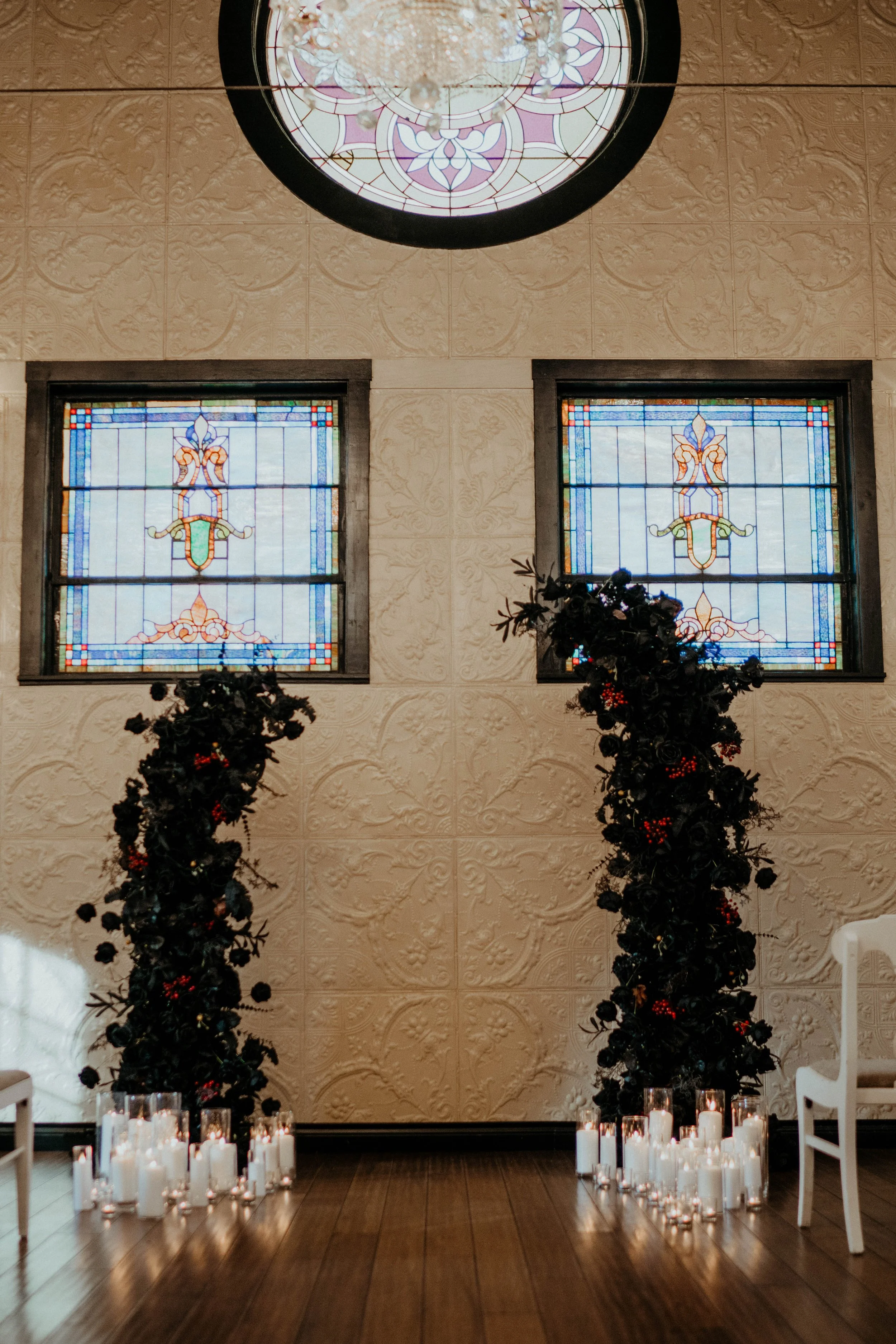 Two black decorated Christmas trees with red ornaments placed in front of a beige textured wall with two stained glass windows, surrounded by white candles on the wooden floor.