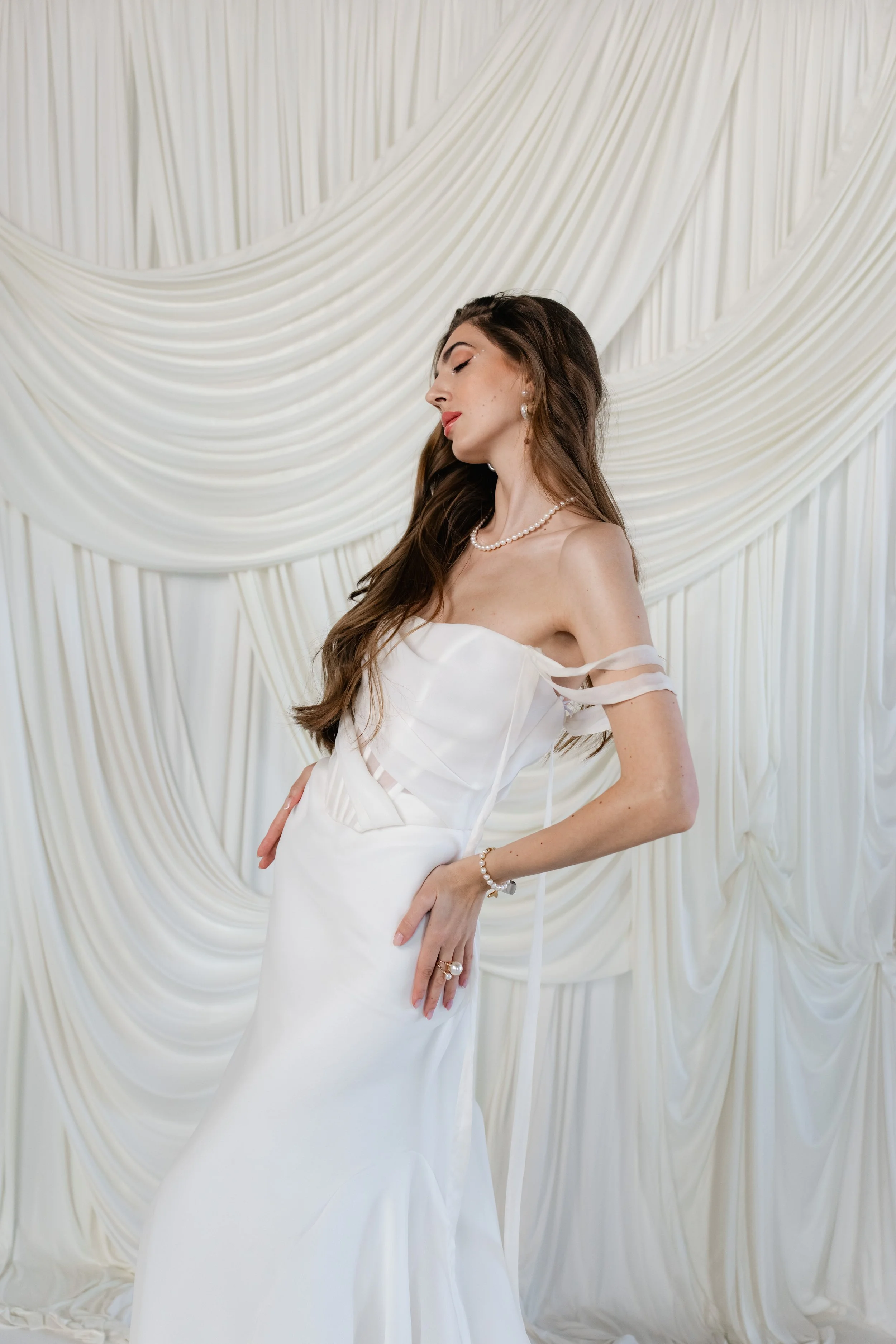 A woman in a white wedding dress with off-the-shoulder ribbon details, standing against a cream-colored, draped fabric backdrop, with her long brown hair cascading over her shoulder, wearing pearl jewelry.