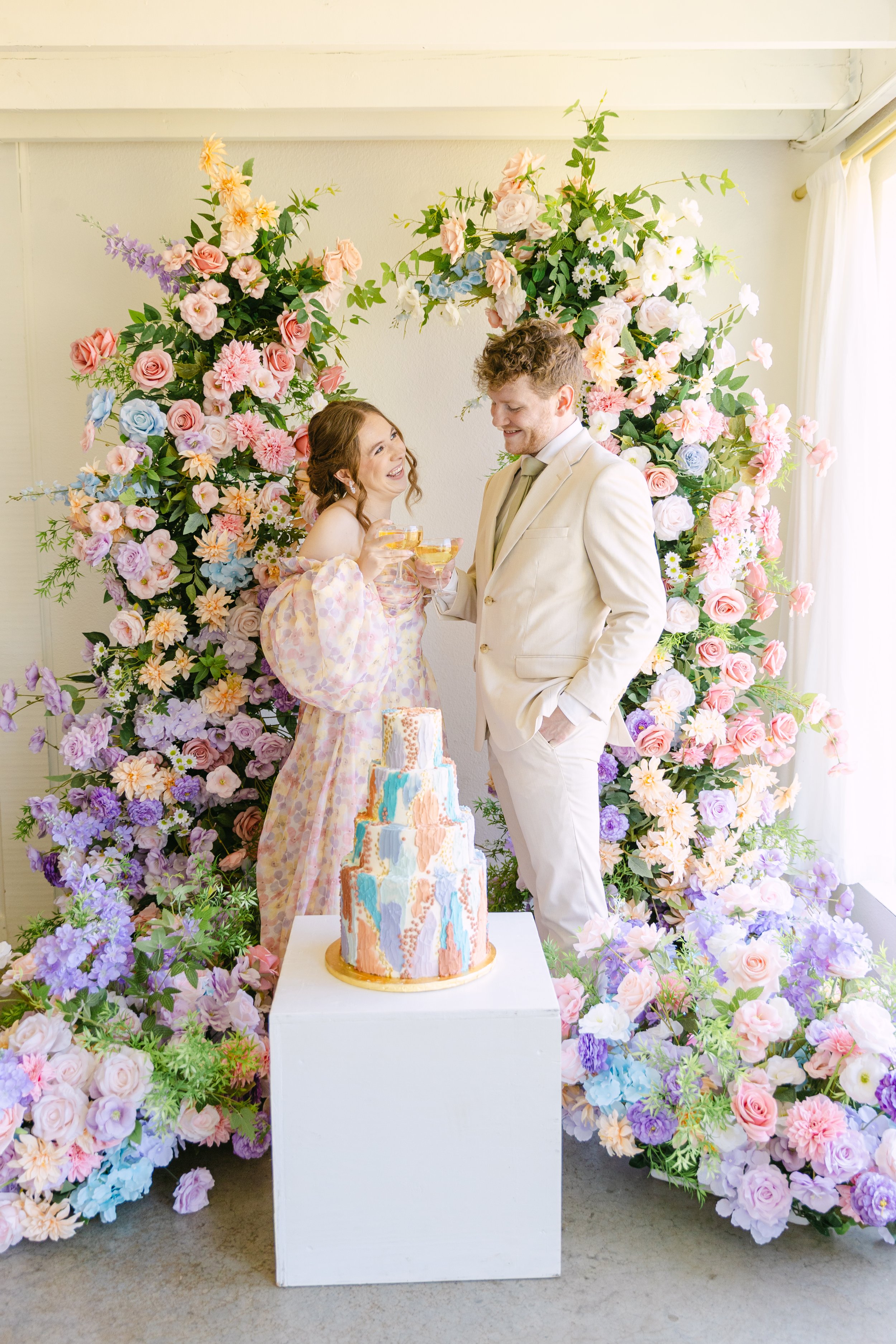 A couple dressed in light-colored formal attire celebrating with champagne in front of a colorful flower backdrop and a tiered cake with pastel-colored icing.