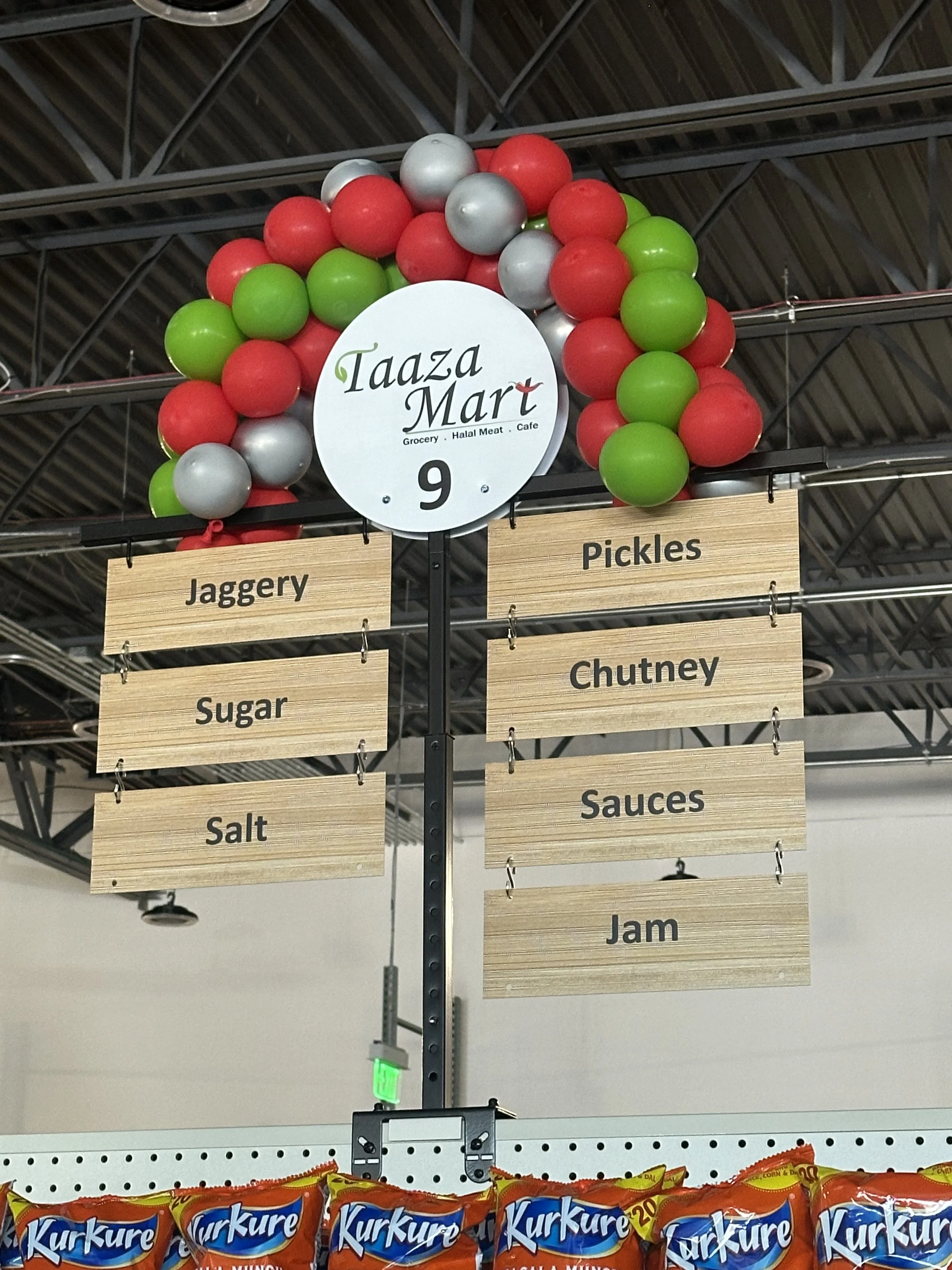 Store sign with balloons reading Taaza Mart, listing products like jaggery, sugar, salt, pickles, chutney, sauces, and jam.