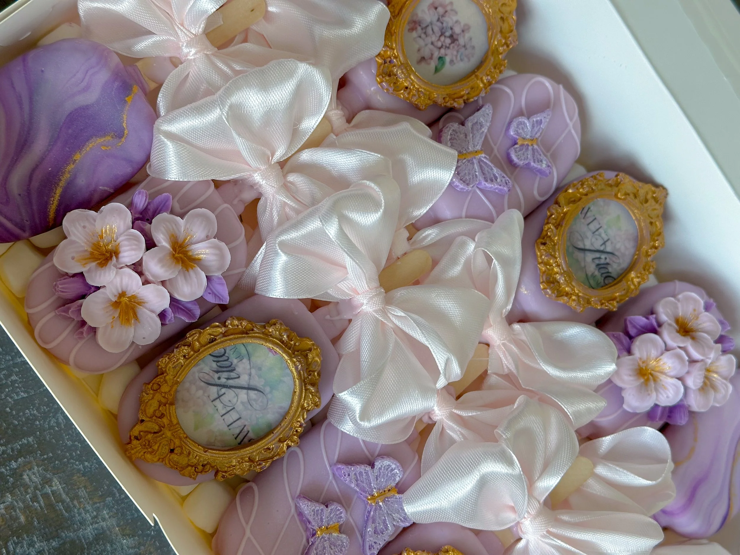 Decorative cakesicles in pastel purple, pink, and yellow with ribbons, flowers, butterflies, and gold-framed edible images.