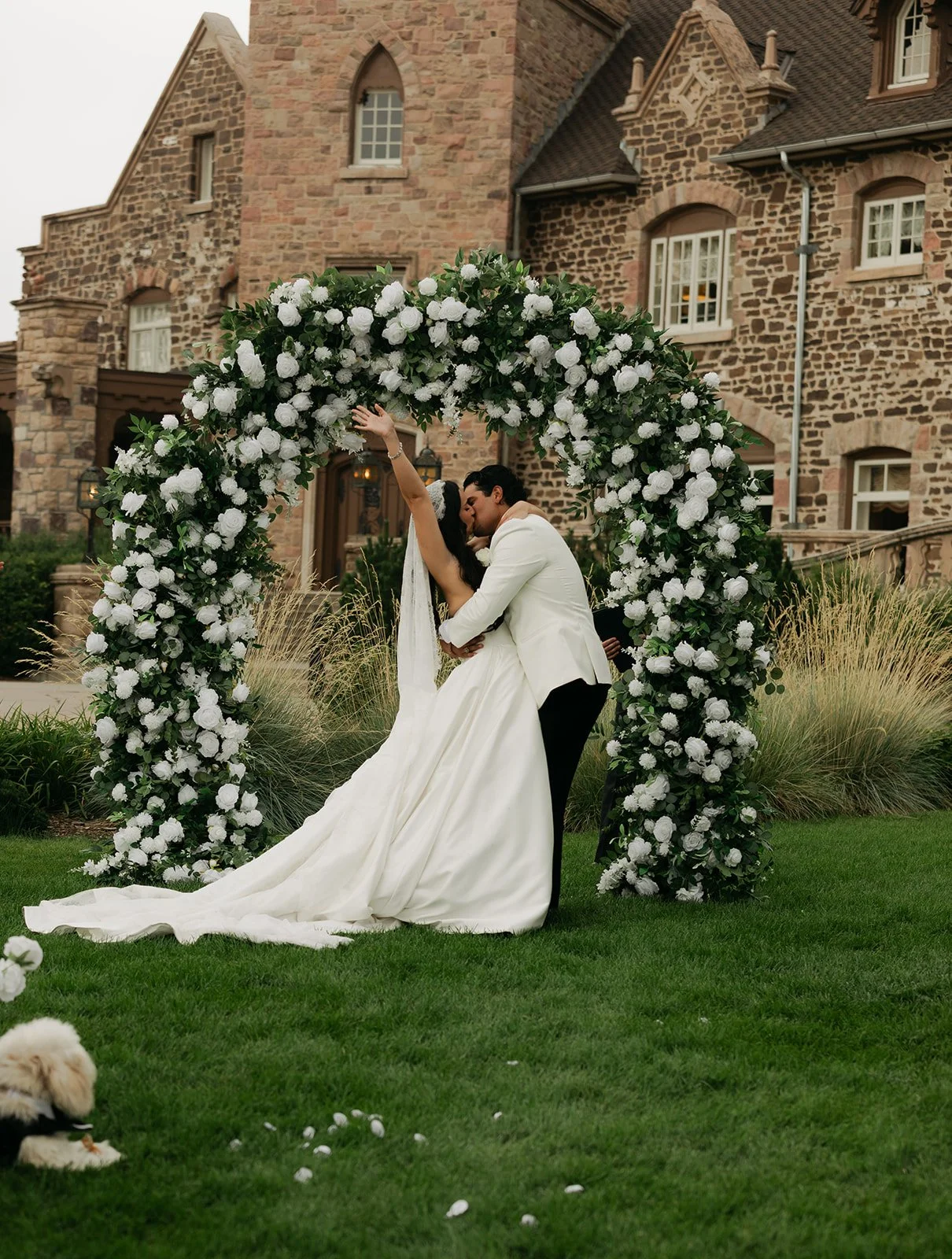A bride and groom share a kiss under a large floral arch outdoors in front of a stone building, with a small dog on the grass nearby.