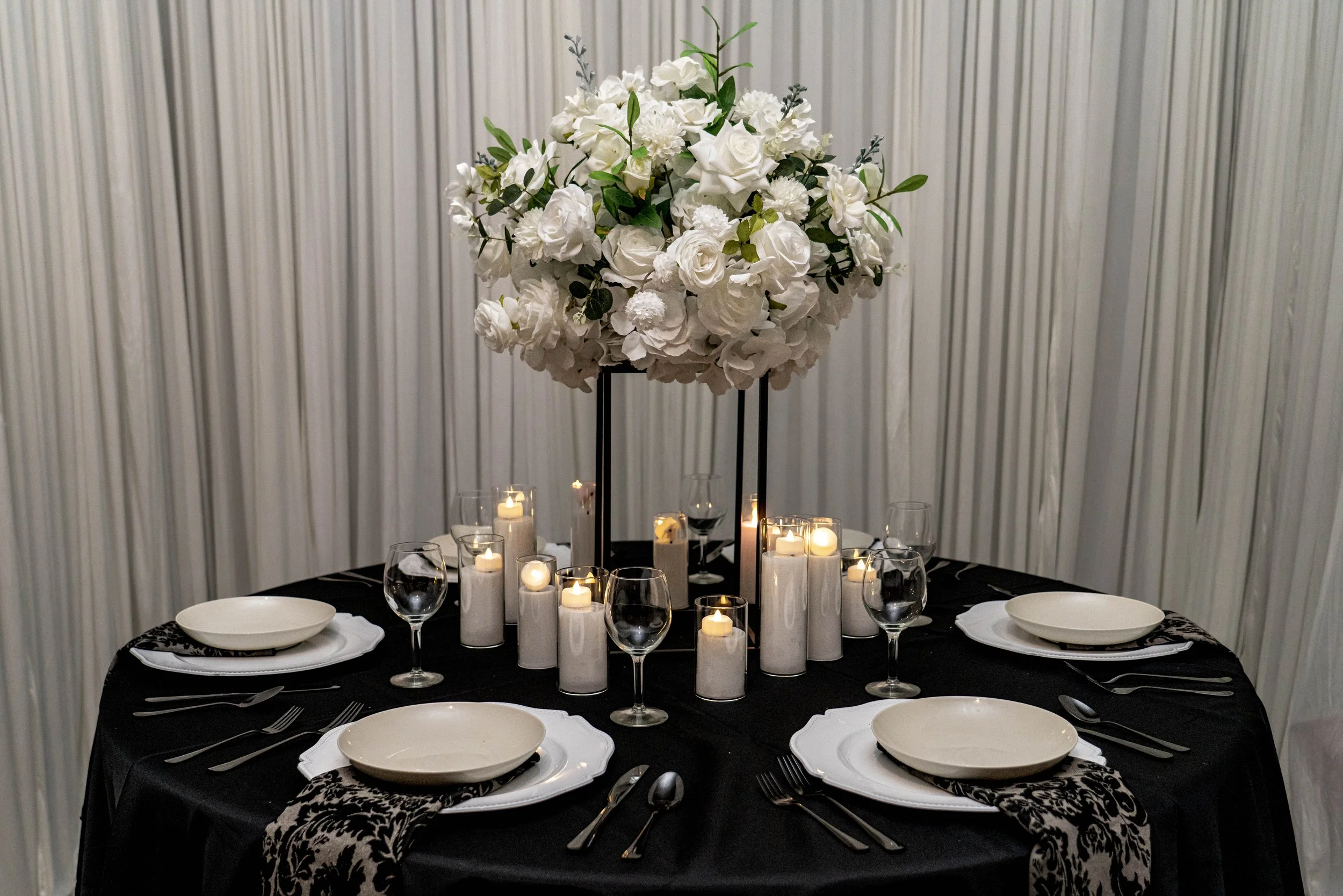 Elegant table setting with white plates, wine glasses, and cutlery, centered around a tall floral arrangement of white flowers and greenery, with lit candles, against a light-colored, vertically-striped wall.