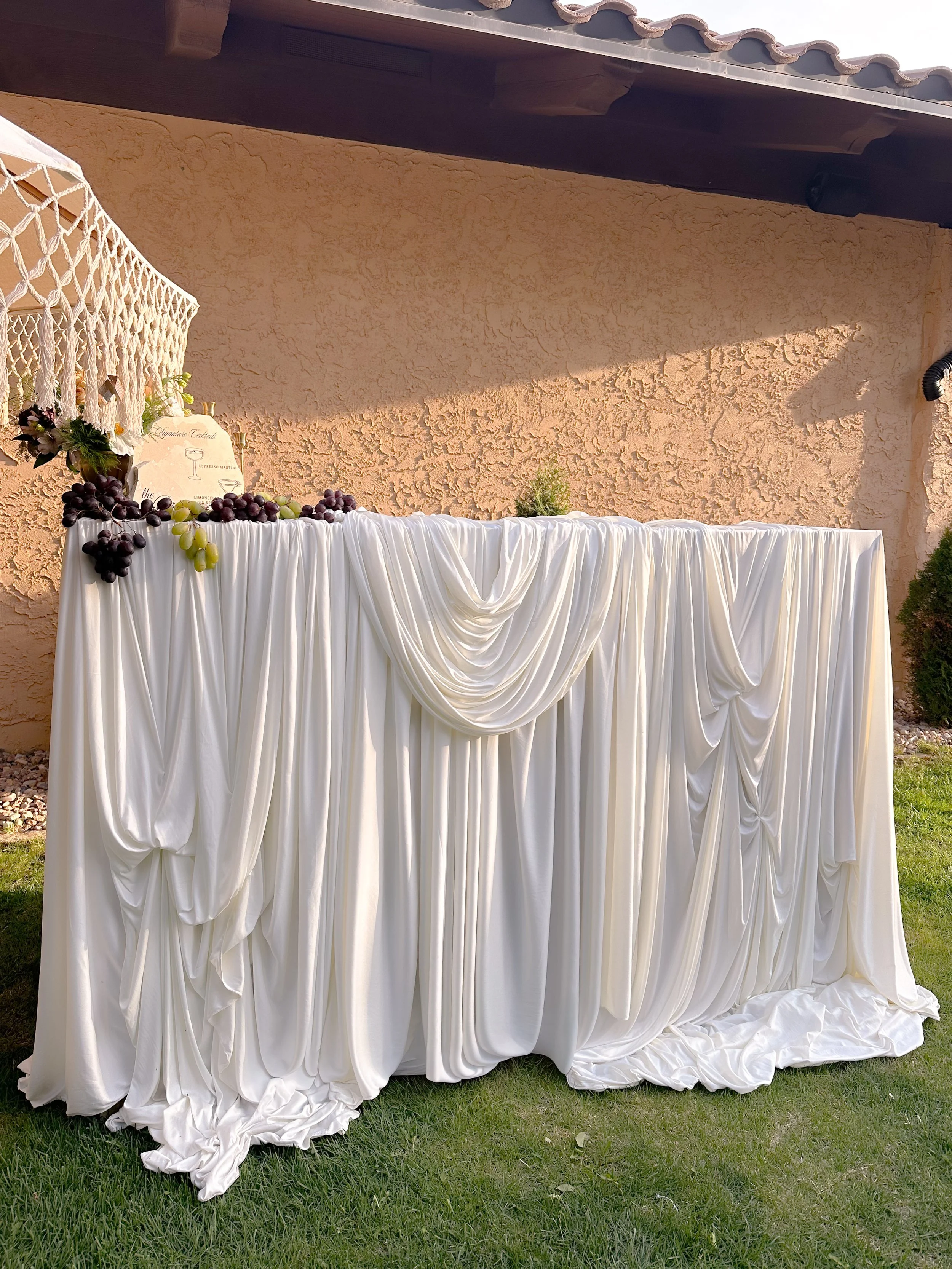 White draped table with grapes and a macrame hanging decoration, set against a textured stucco wall outdoors.