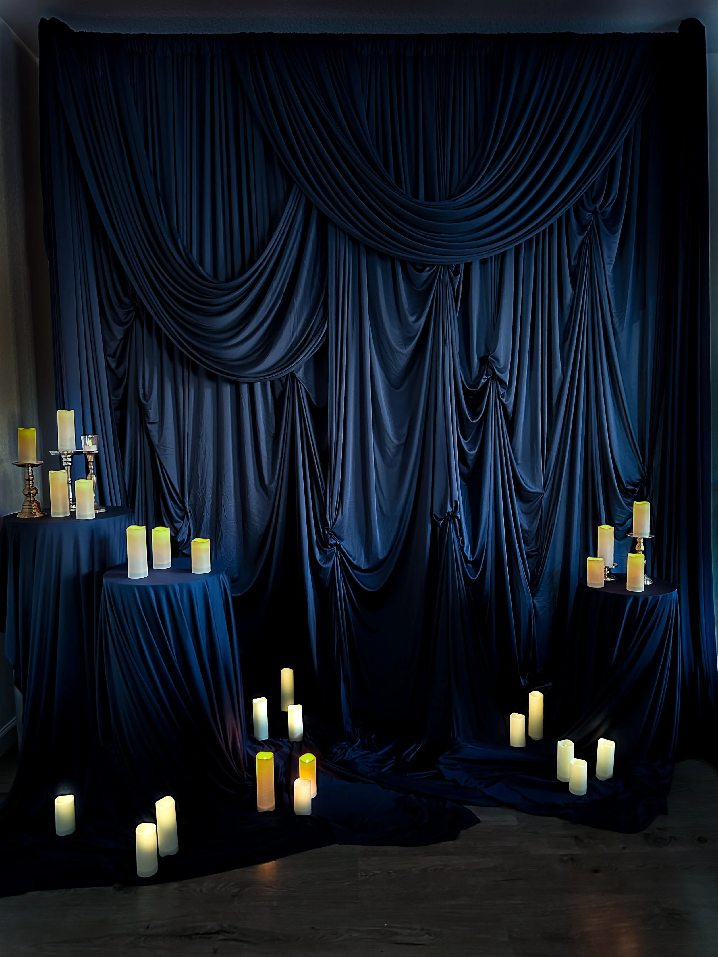 Dark blue draped fabric backdrop with candles on tables and floor, creating an elegant, moody setting suitable for a romantic or mysterious event.
