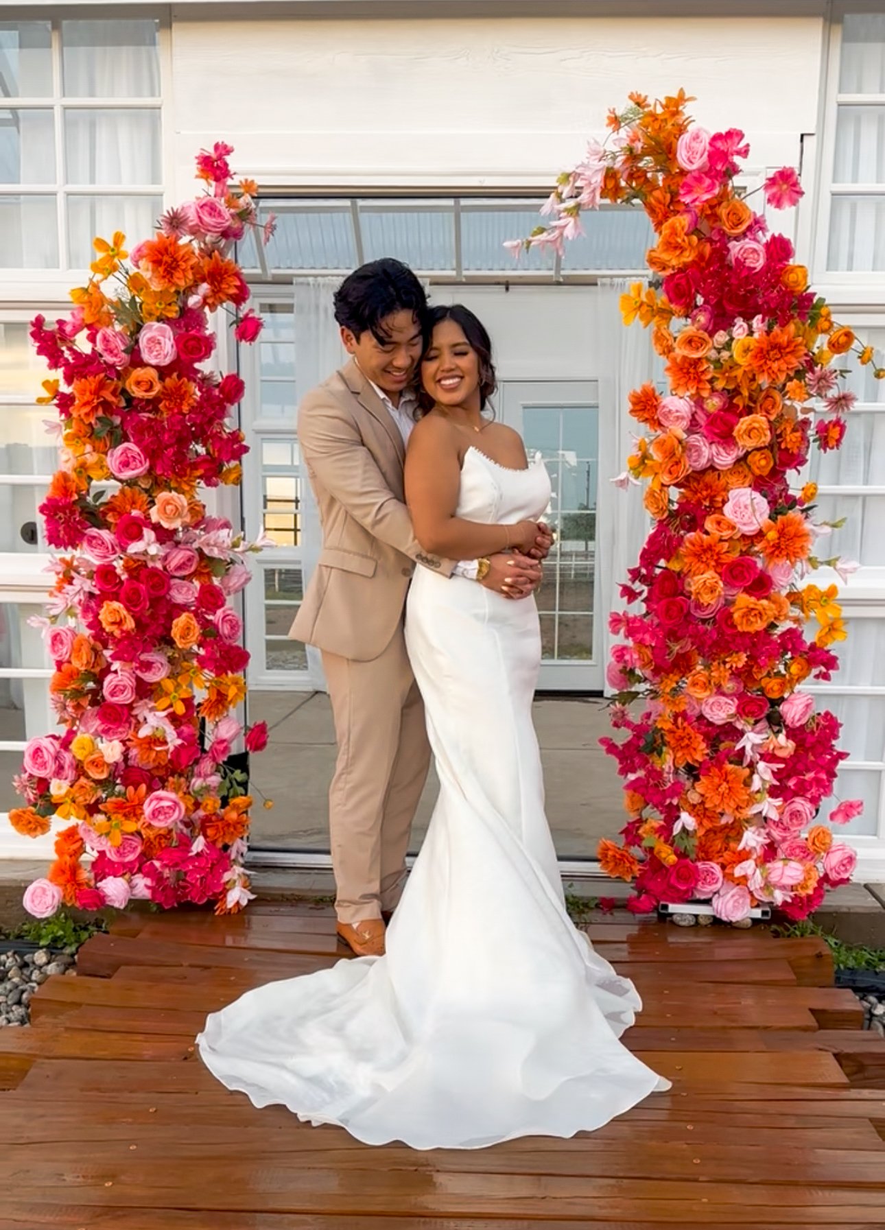 A couple dressed in wedding attire stands in front of a colorful floral arch, with the groom in a beige suit and the bride in a white wedding gown, smiling and embracing each other.