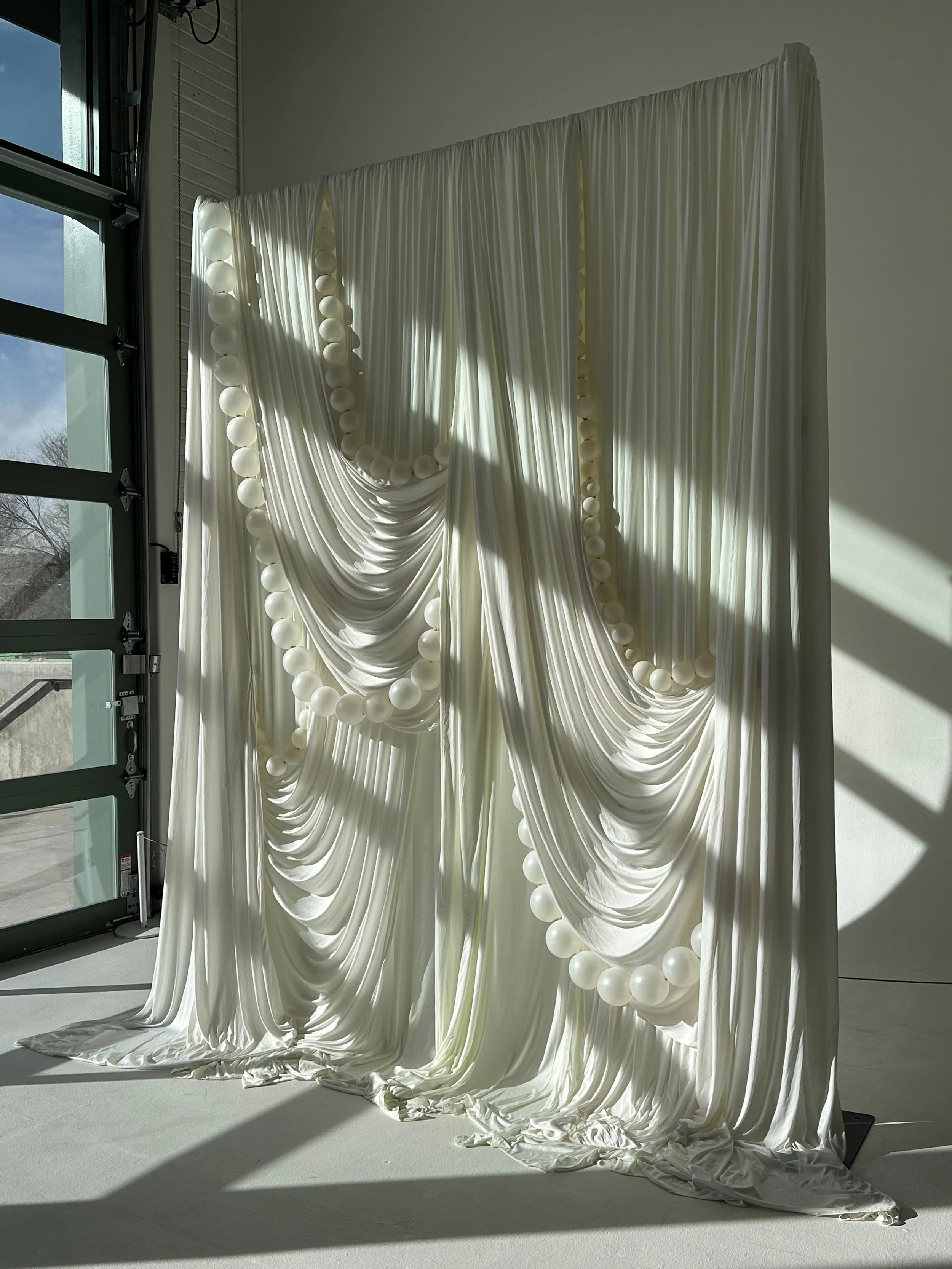 White fabric drapes decorated with multiple strands of white balloons, hanging inside near a large window with sunlight streaming through.