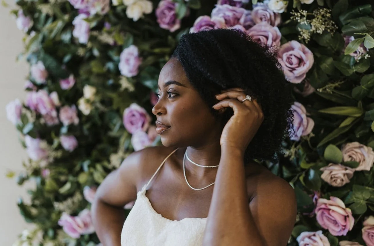 A woman with dark curly hair and dark skin, wearing a white dress and delicate silver necklaces, stands in front of a wall covered with pink and purple roses, touching her hair and looking to the left.