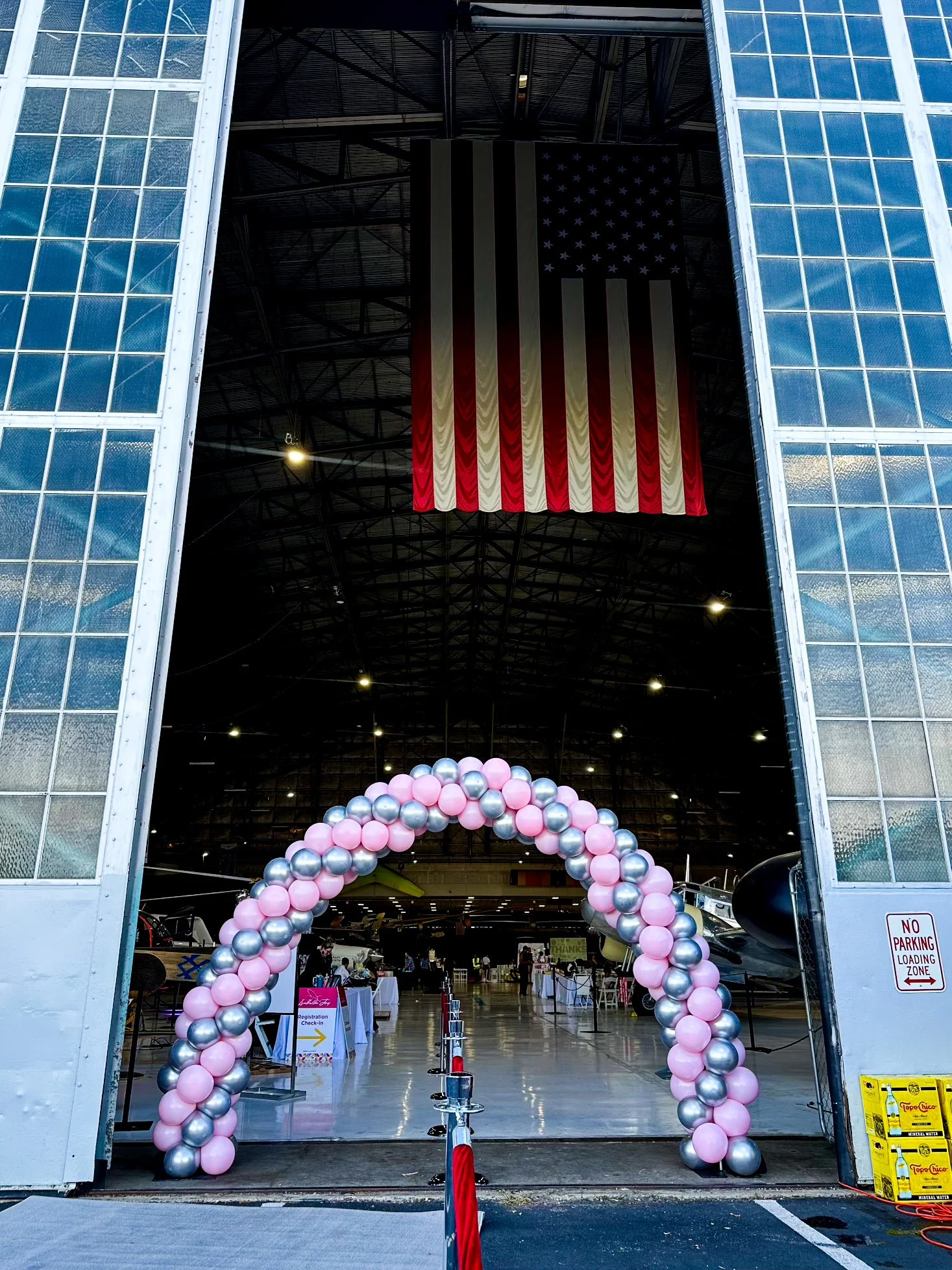 Entrance to an aircraft hangar decorated with pink and silver balloons forming an archway, with a large American flag hanging inside and an American flag banner inside the hangar.