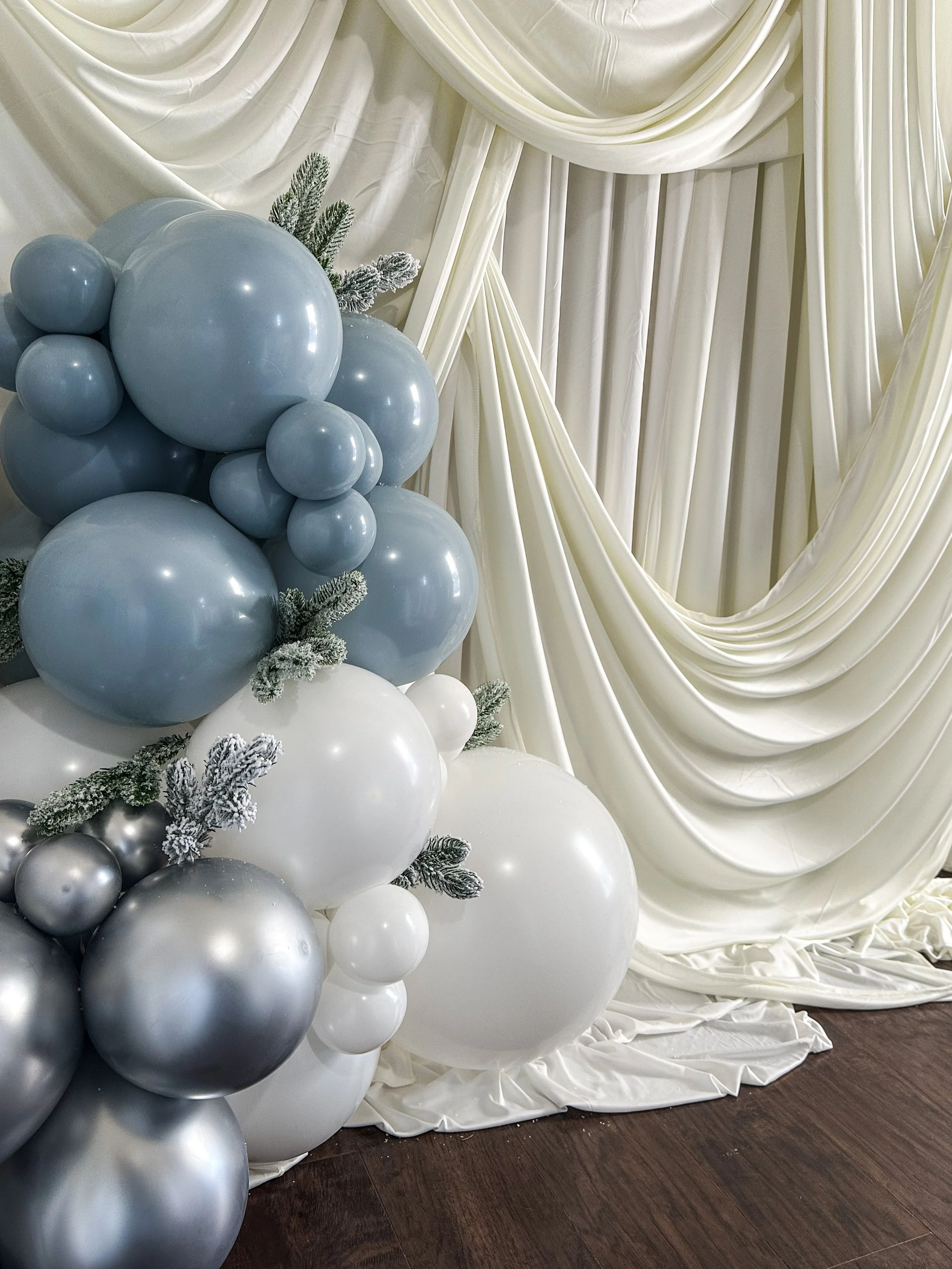 Decorative balloon arrangement with blue, white, and silver balloons, frosted pine branches, and white curtains in the background.