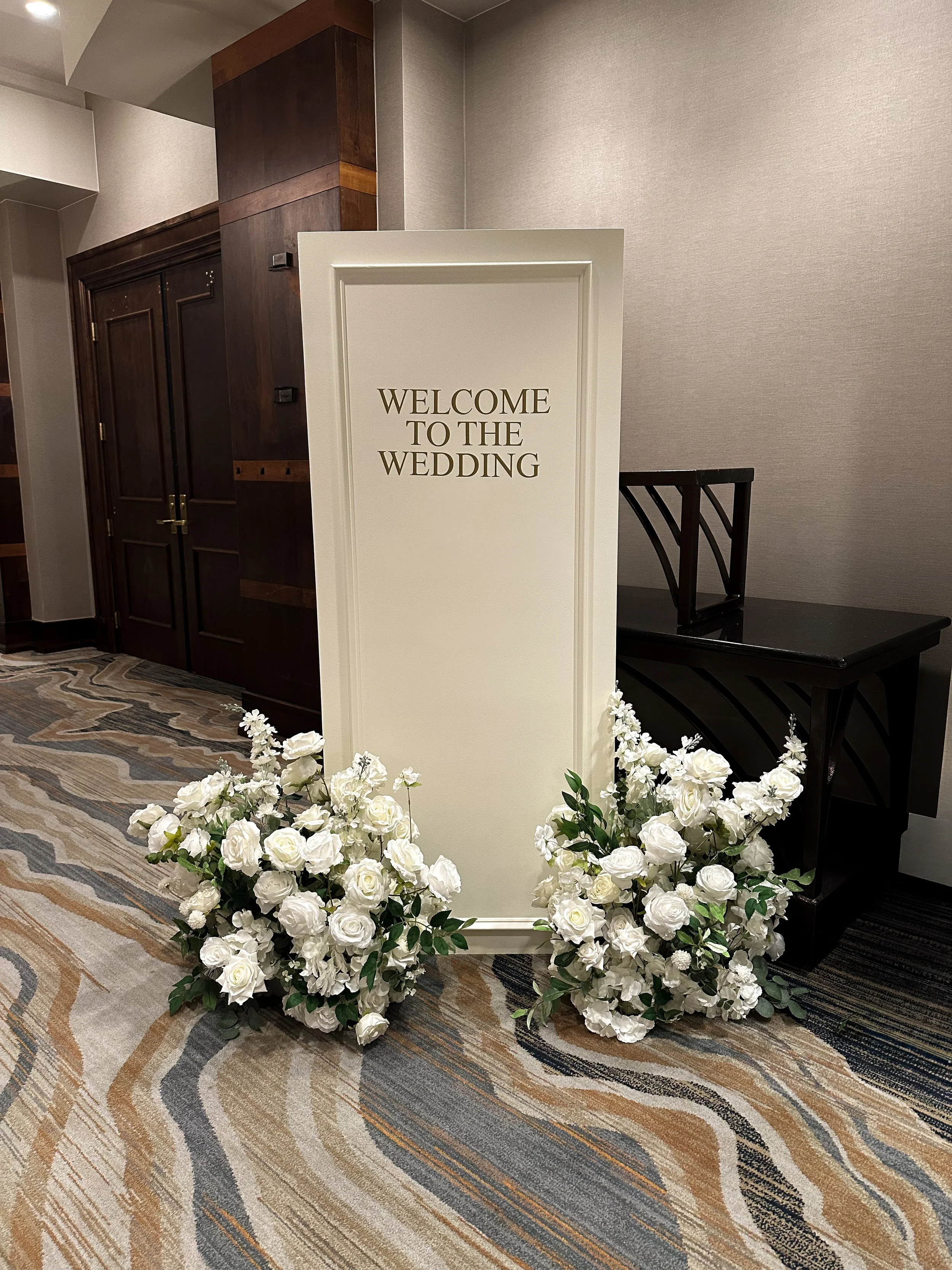 A white sign with gold letters saying 'Welcome to the Wedding', decorated with white flowers at the base, placed in an indoor setting with a carpeted floor, a wooden cabinet, and grey walls.