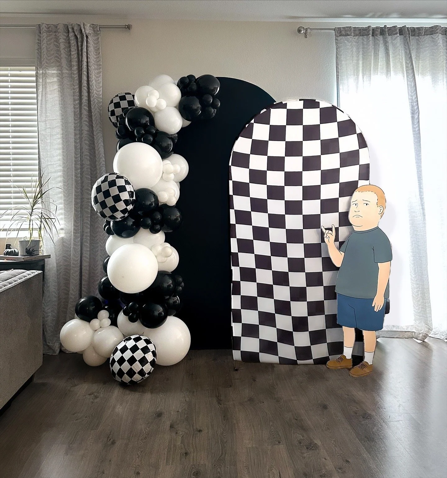 Black and white balloon arch with checkered pattern along one side, decorated backdrop with black, white, and checkered design, and a cardboard cutout of a cartoon man making a rock and roll gesture, inside a room with gray curtains and a wooden floo