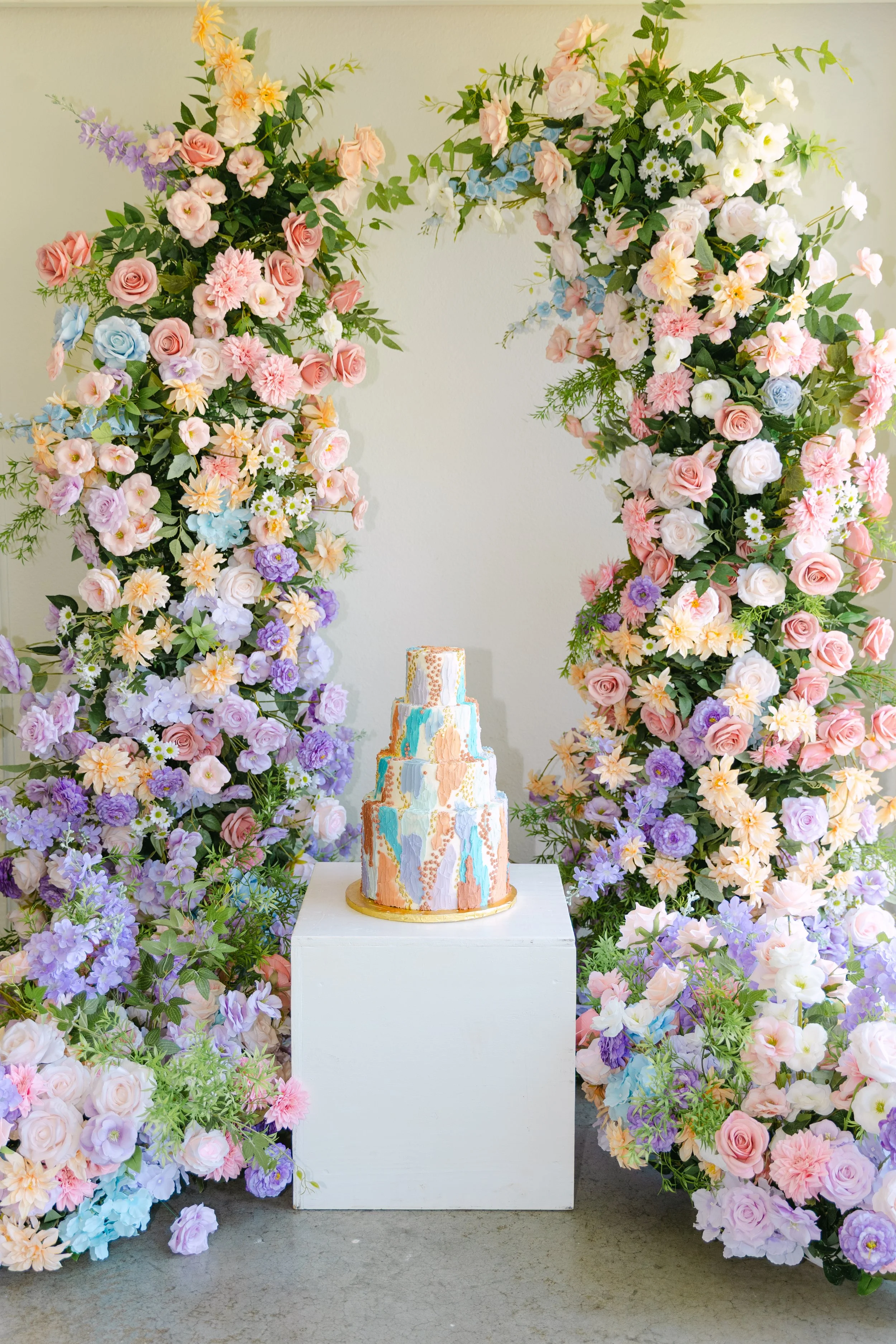 A pastel-colored, abstract, multi-tiered wedding cake is displayed on a white pedestal, surrounded by a large, ornate floral arch with pink, purple, white, and blue flowers and green foliage.