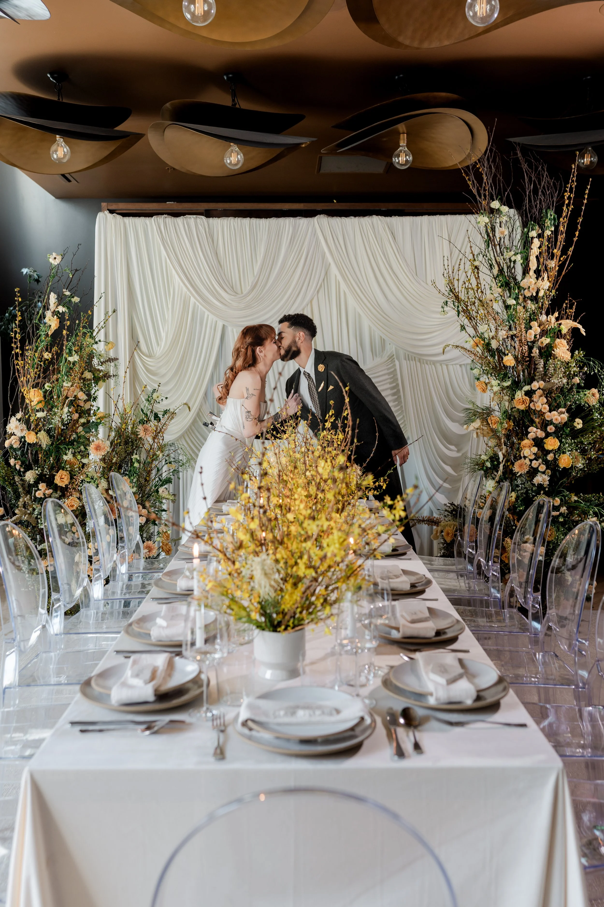 A couple is kissing at a wedding reception table, decorated with flowers and set for a meal, with a white draped backdrop behind them.