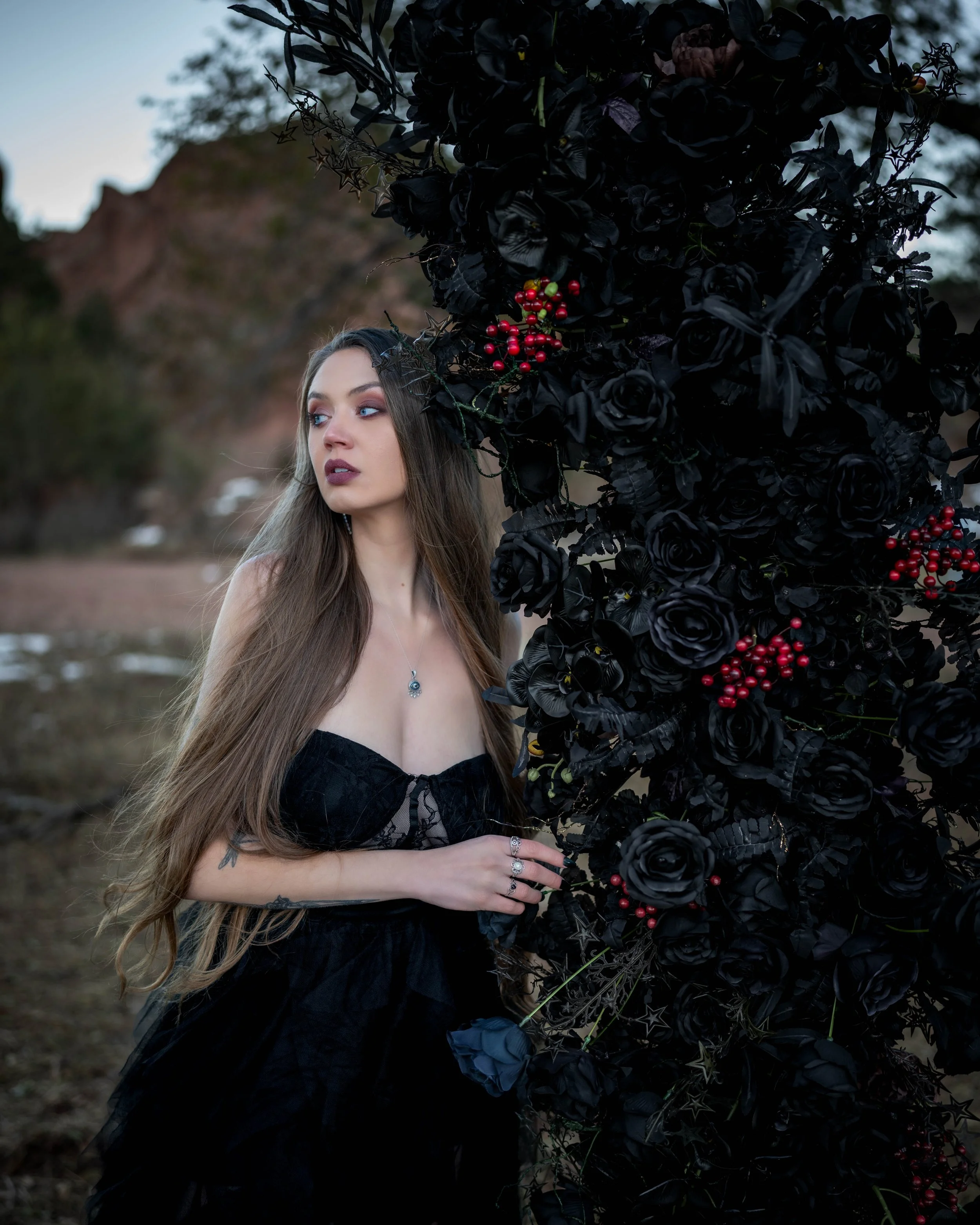 A woman with long brown hair wearing a black lace dress standing next to a large, dark floral arrangement with black roses and red berries outdoors.