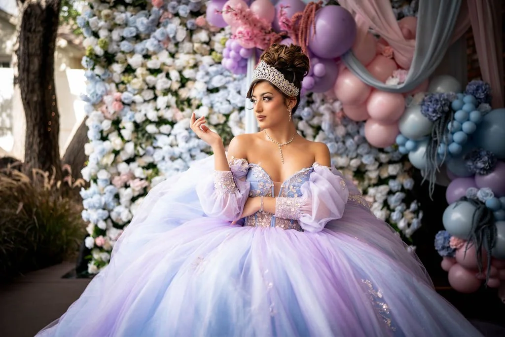 A woman dressed in an elaborate purple ball gown with off-shoulder sleeves, adorned with sequins and lace, sitting in front of a floral backdrop with pink, purple, and white balloons and flowers. She is wearing a tiara and jewelry, with her hair styl