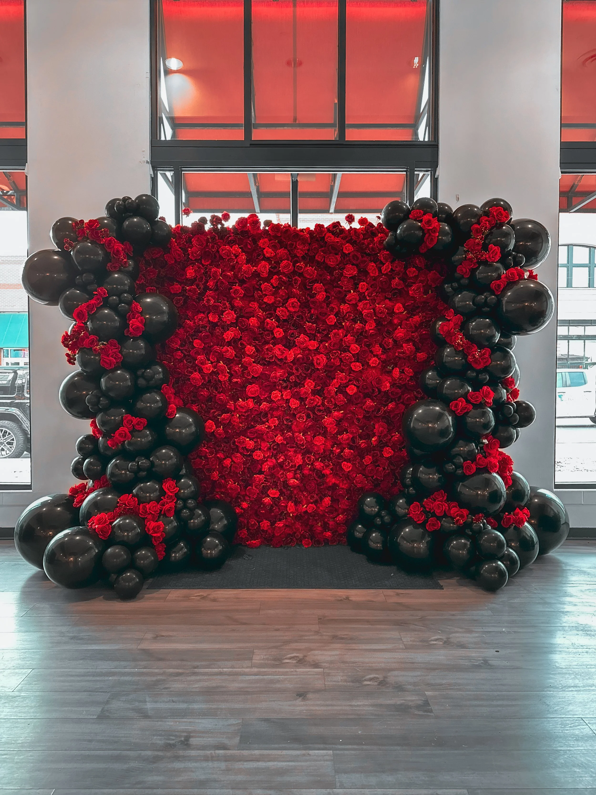 A decorative red rose wall with black balloons surrounding it, placed in front of large windows with a view of parked cars outside.