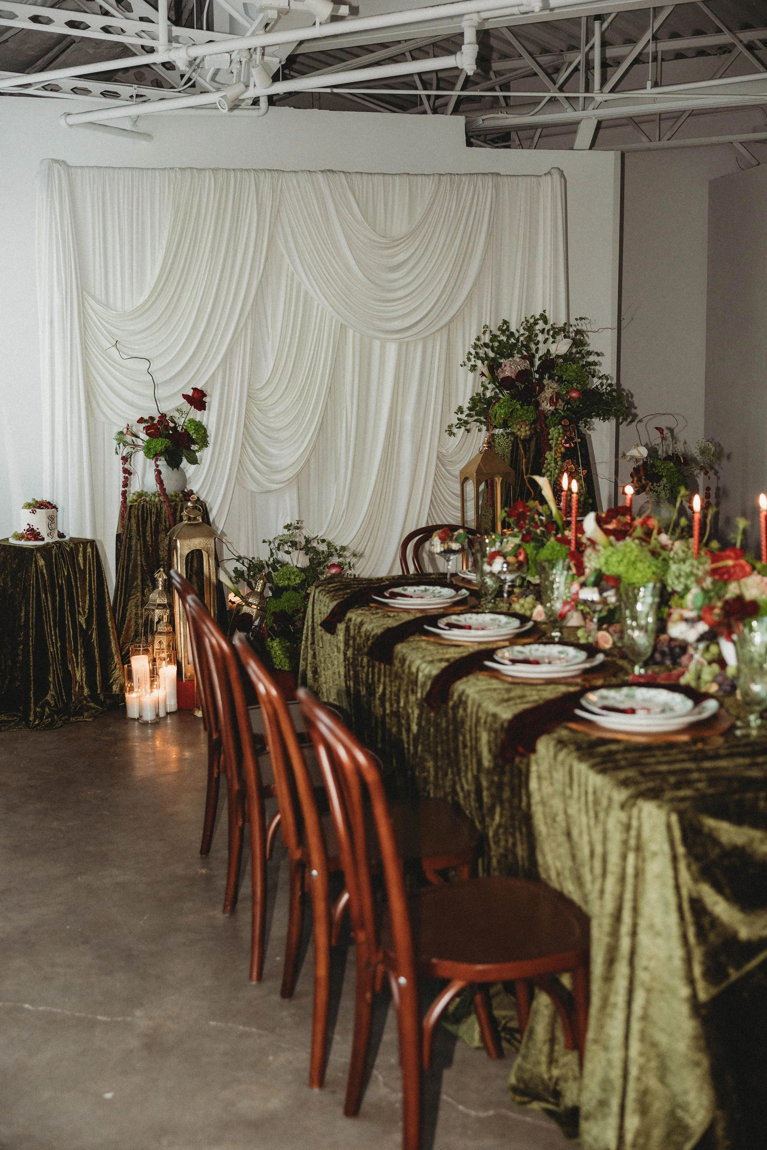Elegant banquet table decorated with floral centerpieces, candles, and gold and dark cloth linens, set against a white draped backdrop with flower arrangements and lanterns, in a modern indoor event space.