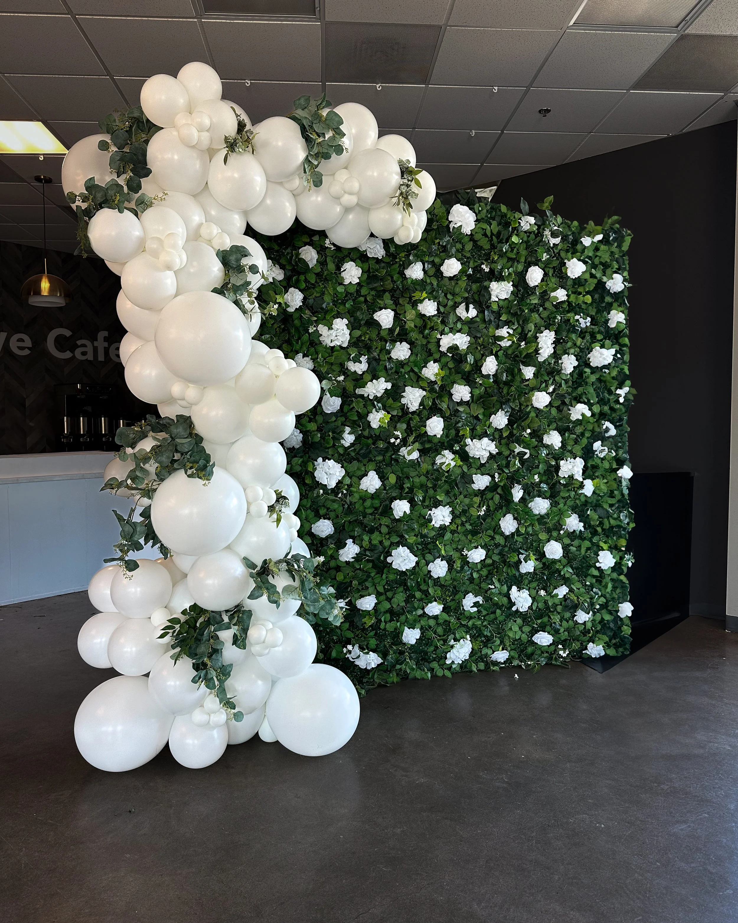 A decorative backdrop with a lush green foliage wall and white flowers, accented with a cluster of white balloons and greenery on the left side.