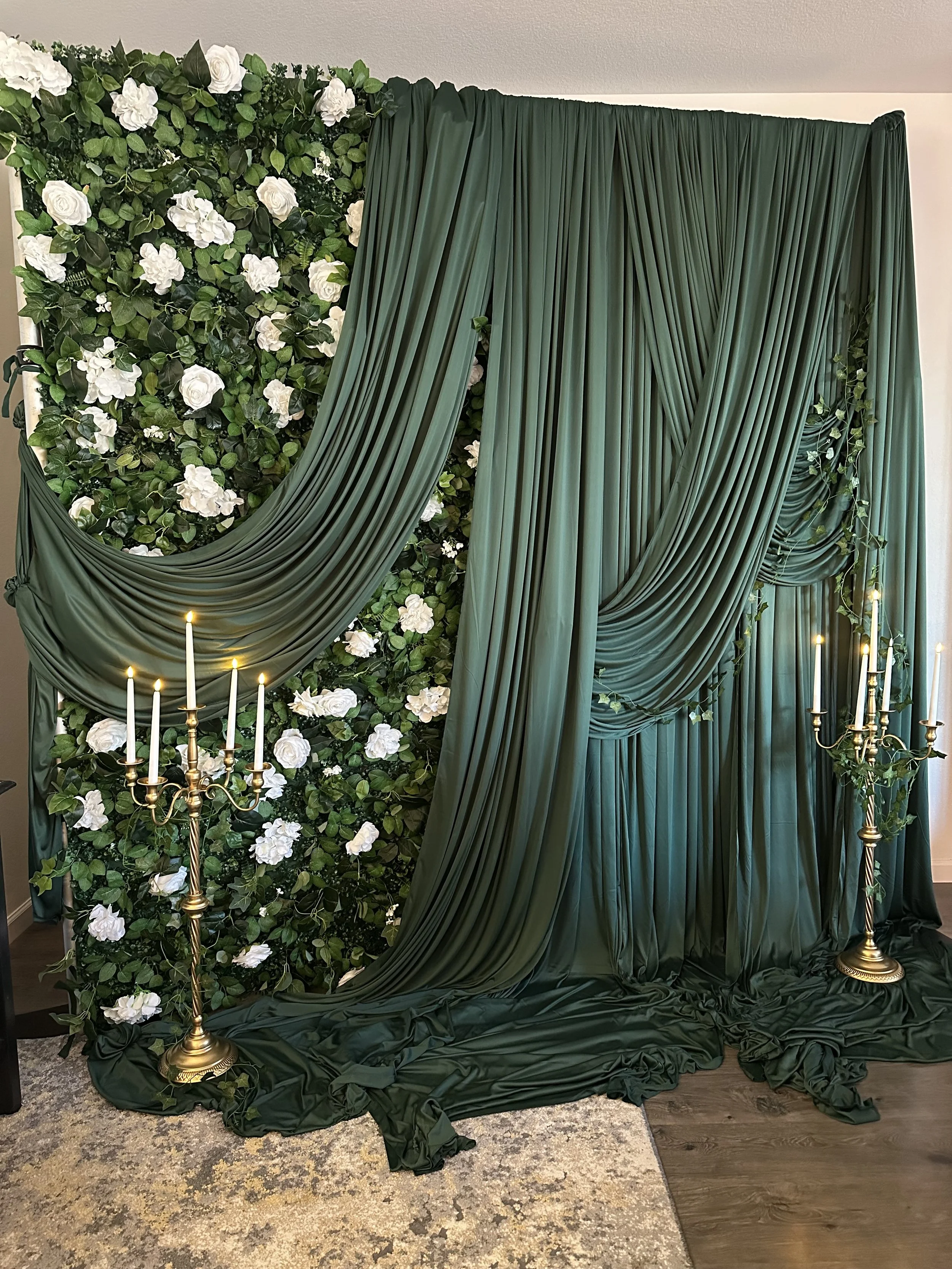 A decorative backdrop with green draped fabric, white flowers, greenery, and two gold candelabras with lit candles.