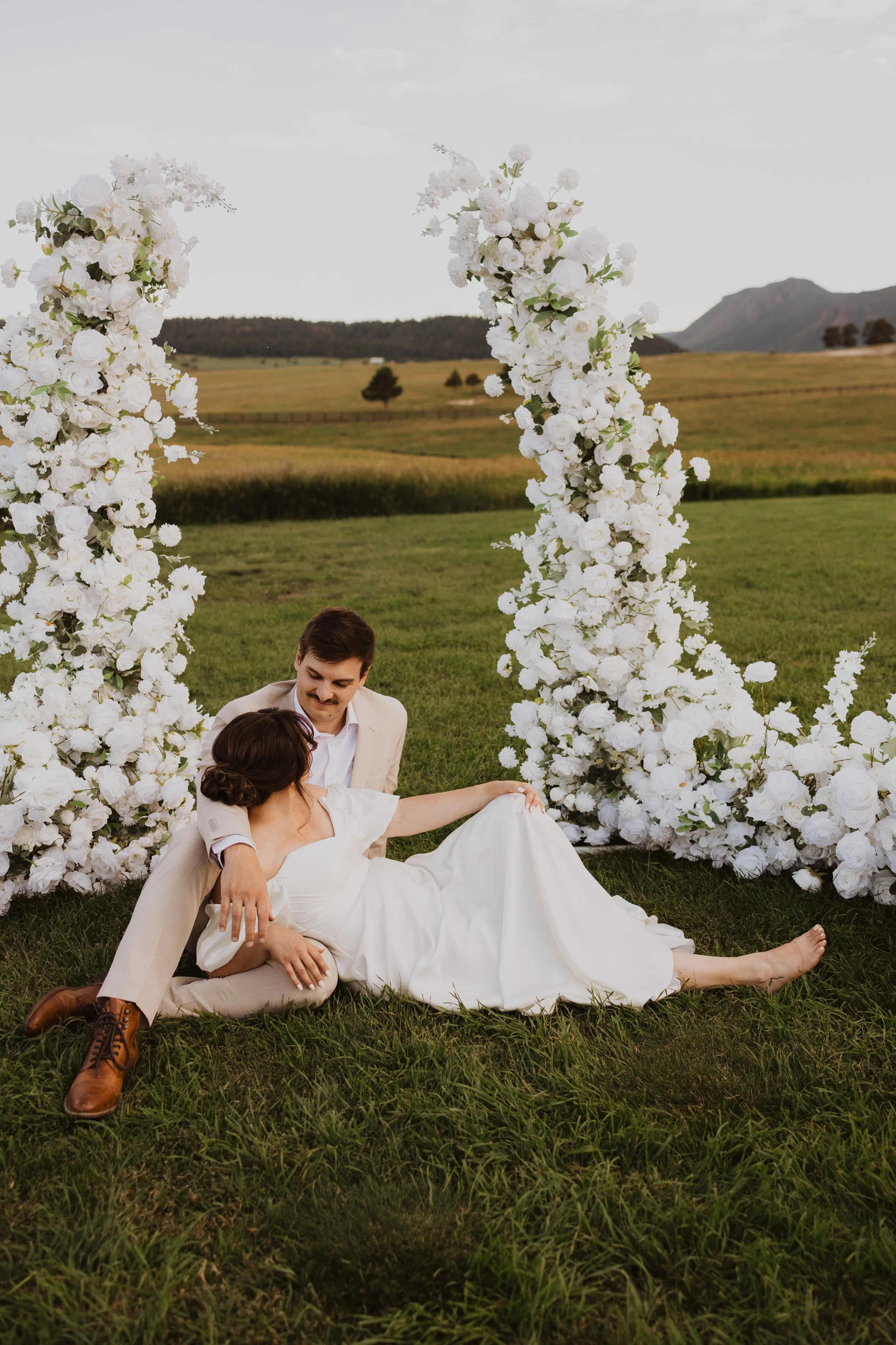 A couple in wedding attire sitting on grass in front of a large white floral arch outdoors, with a scenic field and mountains in the background.