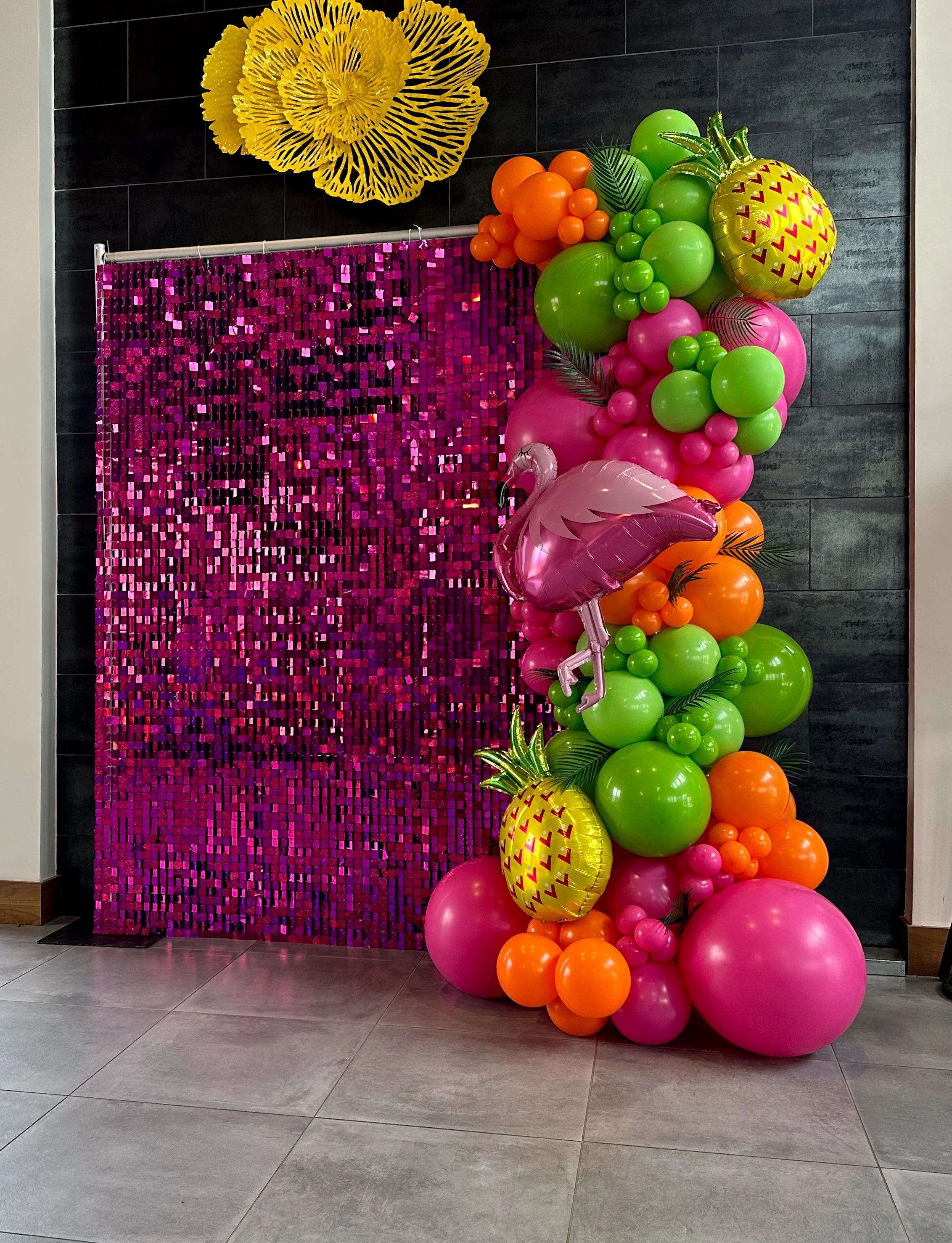 Colorful balloon display featuring pink flamingo, pineapples, and various green, orange, and pink balloons arranged next to a pink sequin backdrop and yellow paper flower decoration on a black tiled wall.