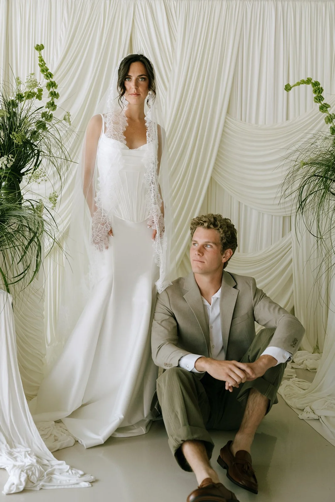 A woman in a wedding dress with a veil stands next to a seated man in a suit, in front of a white drapery backdrop and green plants.