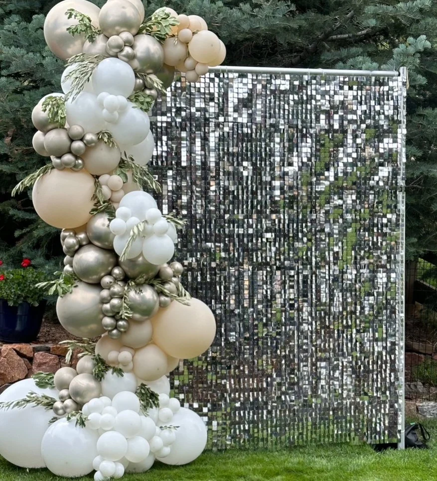 Decorative backdrop with a large, curved balloon arrangement in shades of white, cream, gold, and silver, with greenery, next to a reflective, mirror-like sequin curtain, set outdoors with trees in the background.